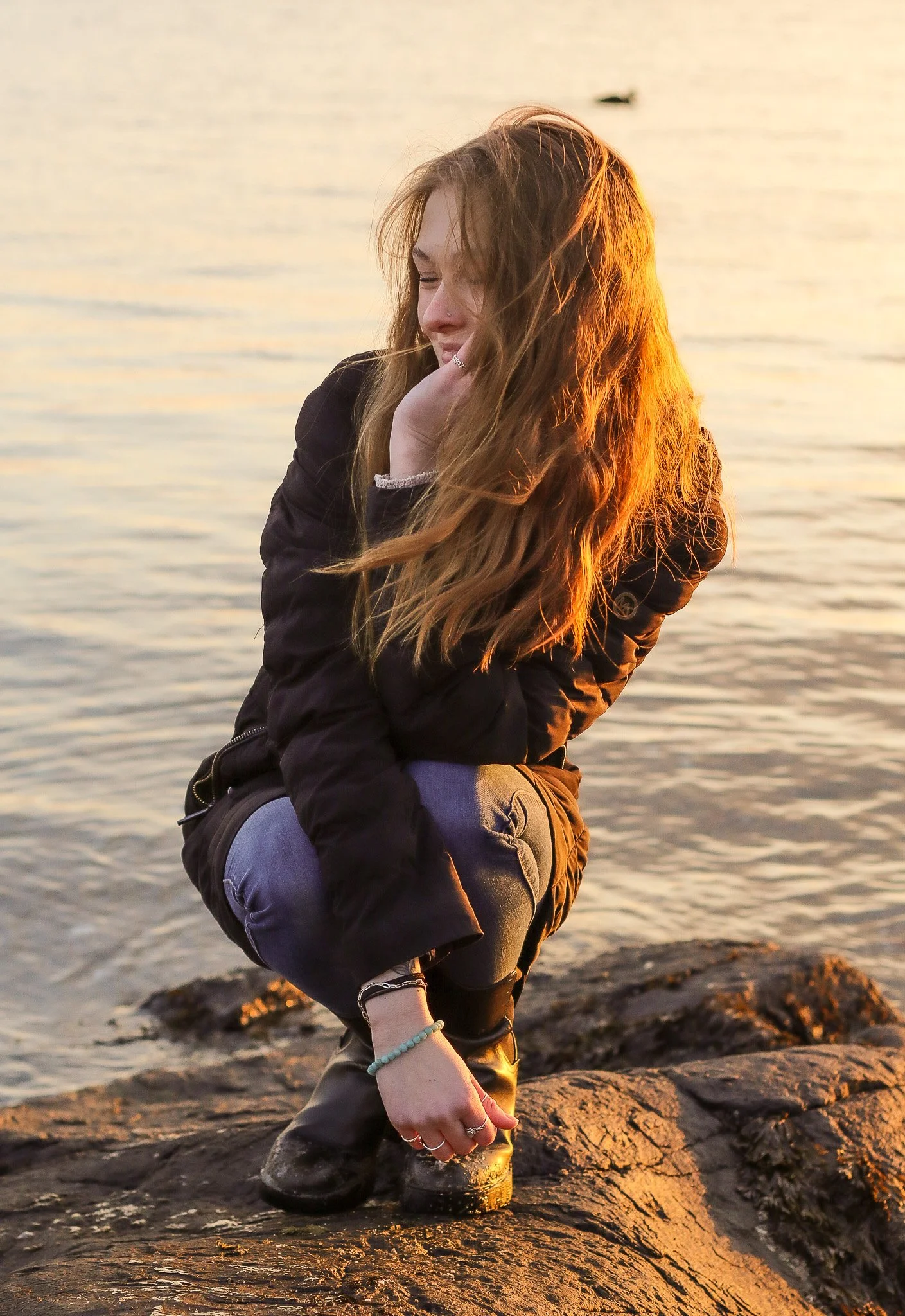 A young woman with long red hair crouching on a large rock by a body of water during sunset, smiling gently with her hand resting on her chin.