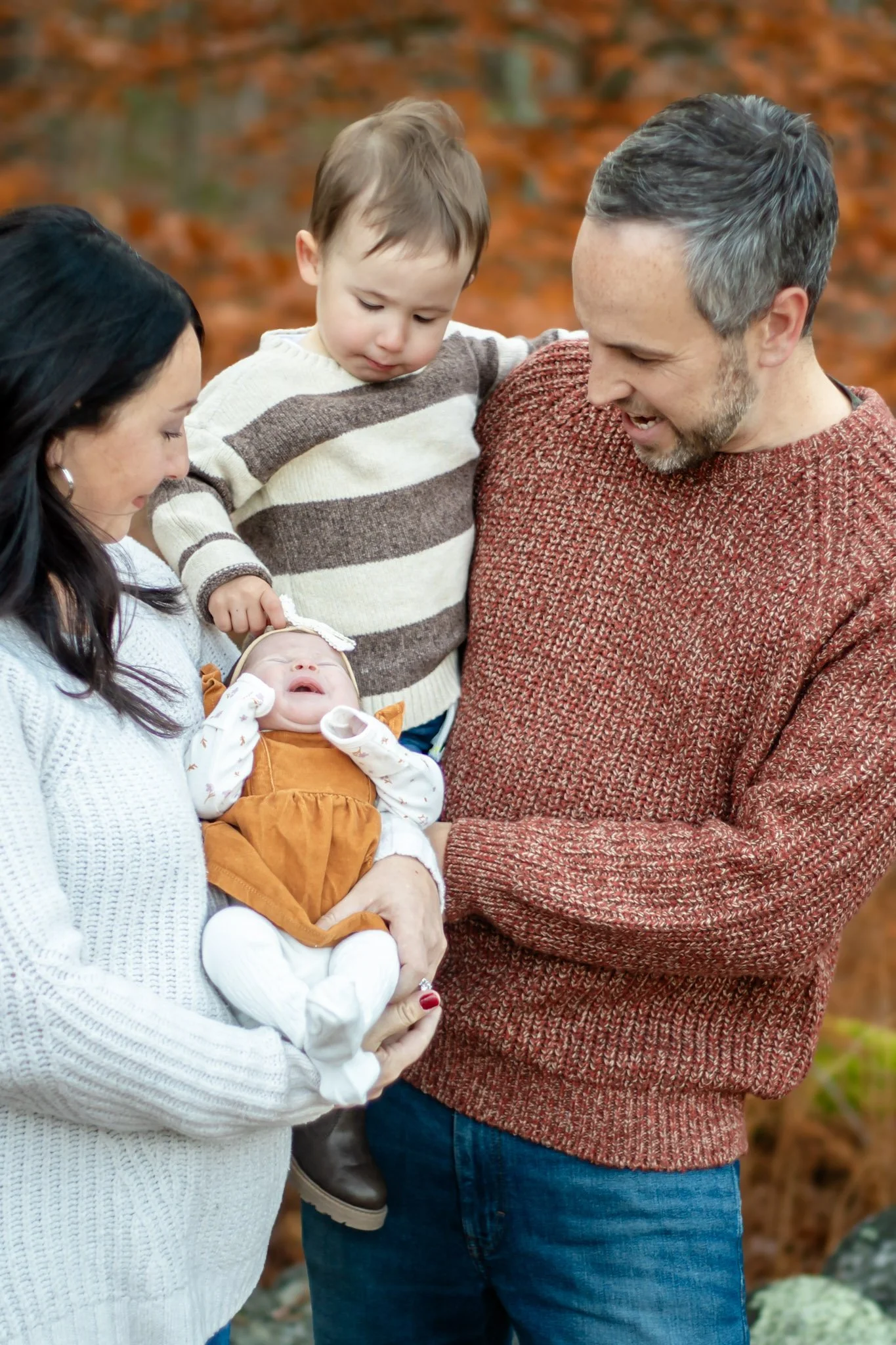 A family of five, including a baby, a toddler, a woman, a man, and another child, enjoy an outdoor fall setting with orange leaves in the background, smiling and engaging with each other.