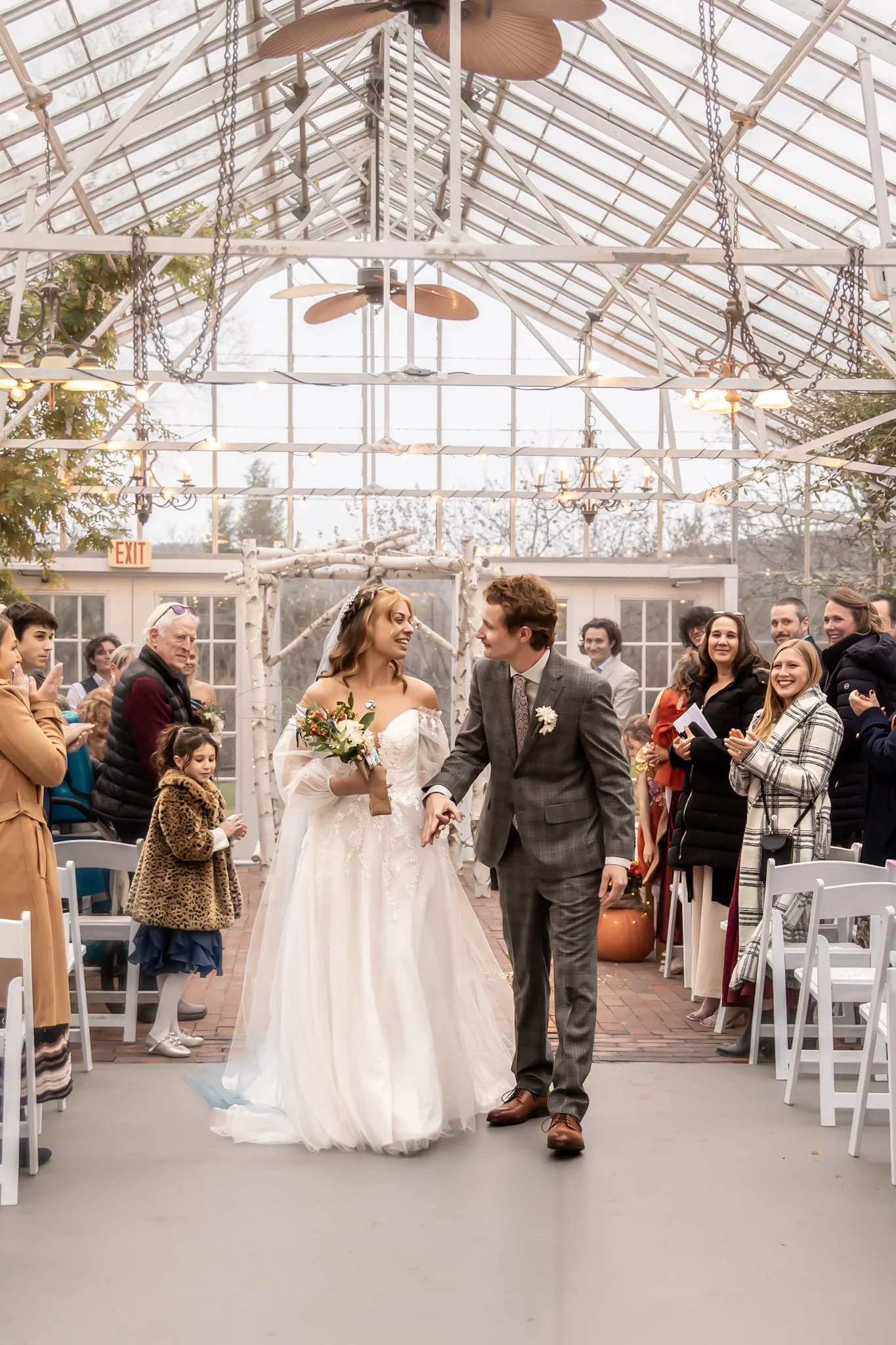 Bride and groom walking down the aisle at a wedding ceremony in a greenhouse with guests clapping and smiling.