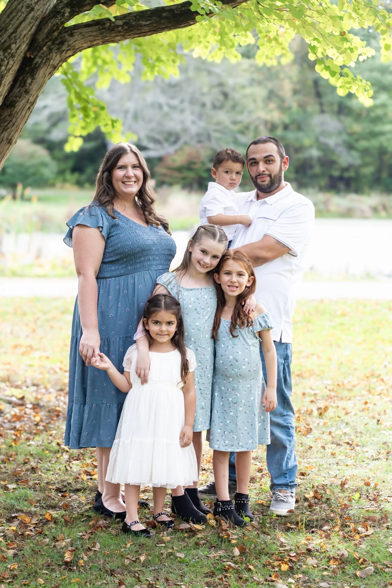 A family of seven standing outdoors under a large tree with green leaves, in a park surrounded by fallen autumn leaves. The group includes a woman in a blue dress, a man in a white shirt holding a young boy, and four young girls in dresses, all smili