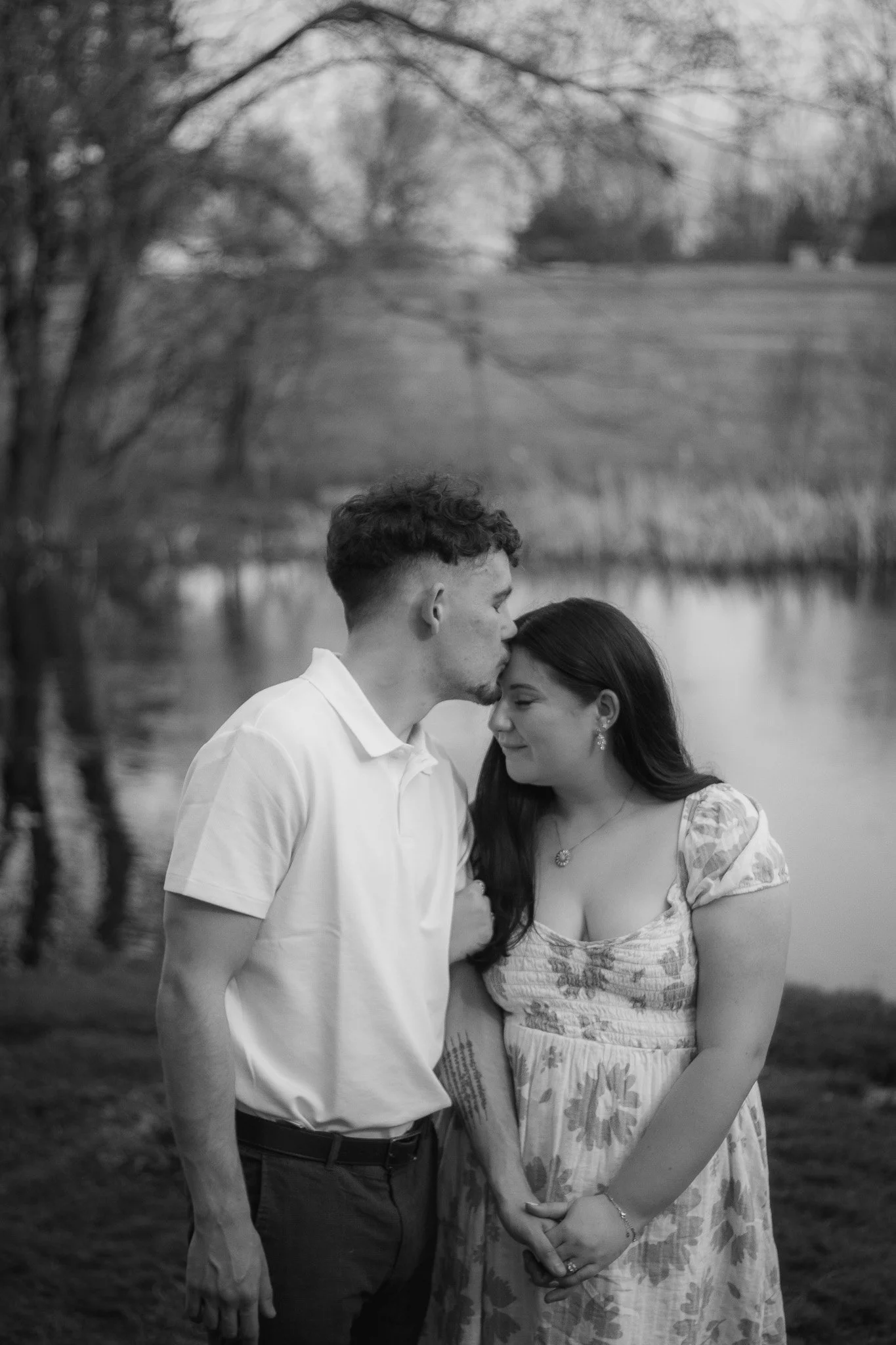 A black and white photo of a couple near a pond, with trees and distant hills in the background. The man is kissing the woman's forehead while she smiles with her eyes closed, holding hands.