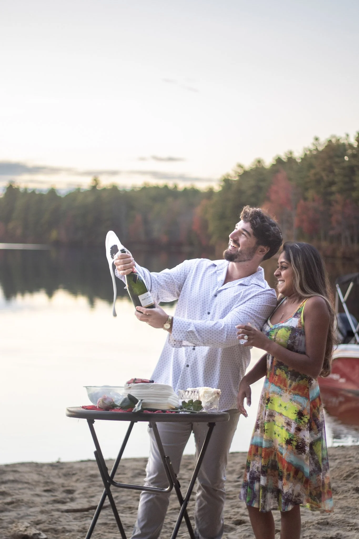 A man and woman celebrating at a lakeside, with a table of cake and food, as the man pops open a bottle of champagne, with a boat in the background and trees on the horizon during sunset.