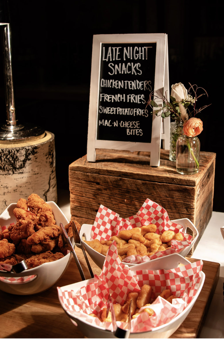 Baskets of fried chicken tenders, French fries, sweet potato fries, and mac and cheese bites on a table with a chalkboard sign listing late night snacks, and a small flower vase with flowers.