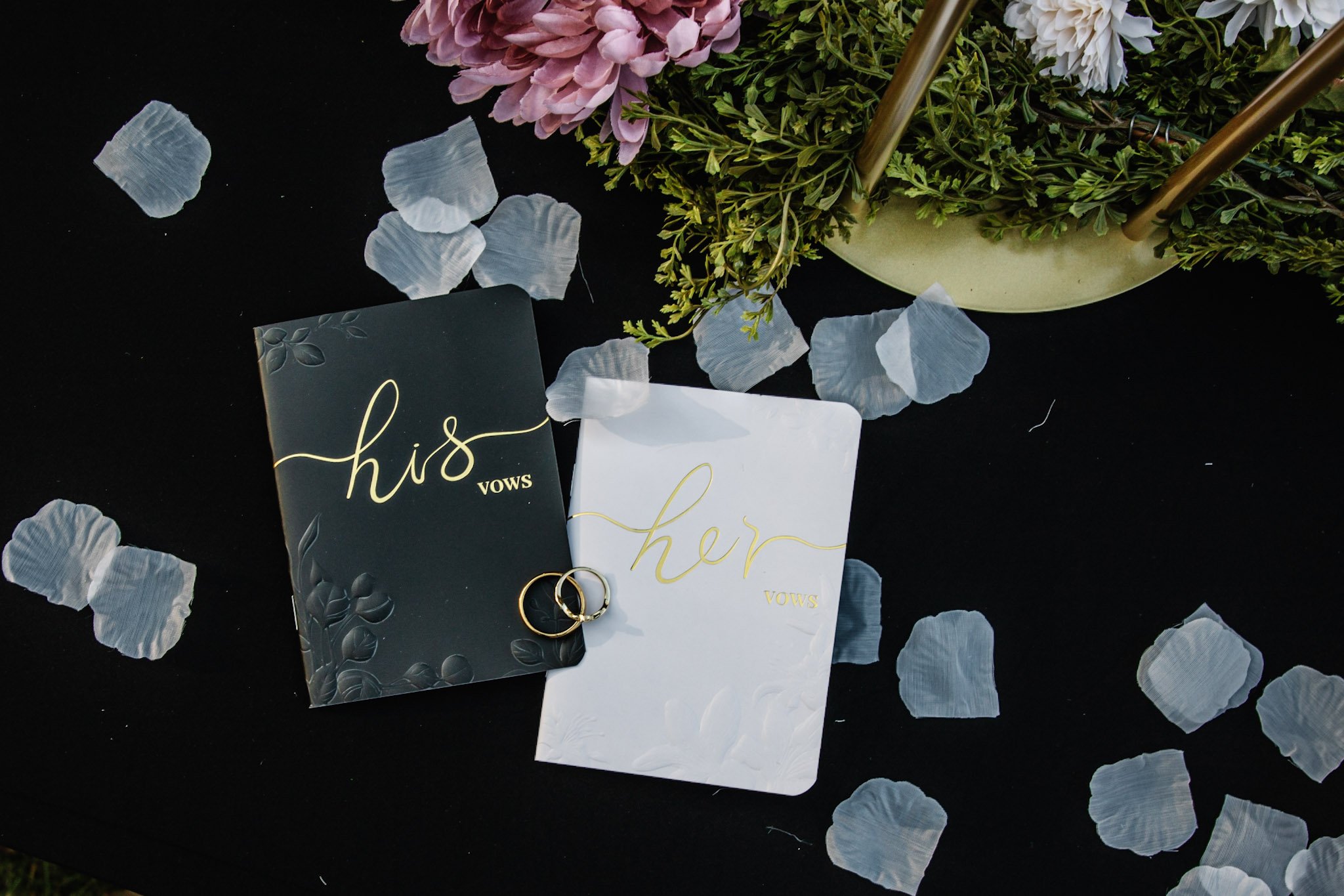 Two vow books labeled "his" and "her" with gold script, positioned on a black surface surrounded by scattered flower petals; a bouquet of pink and white flowers with greenery in a yellow vase in the background.