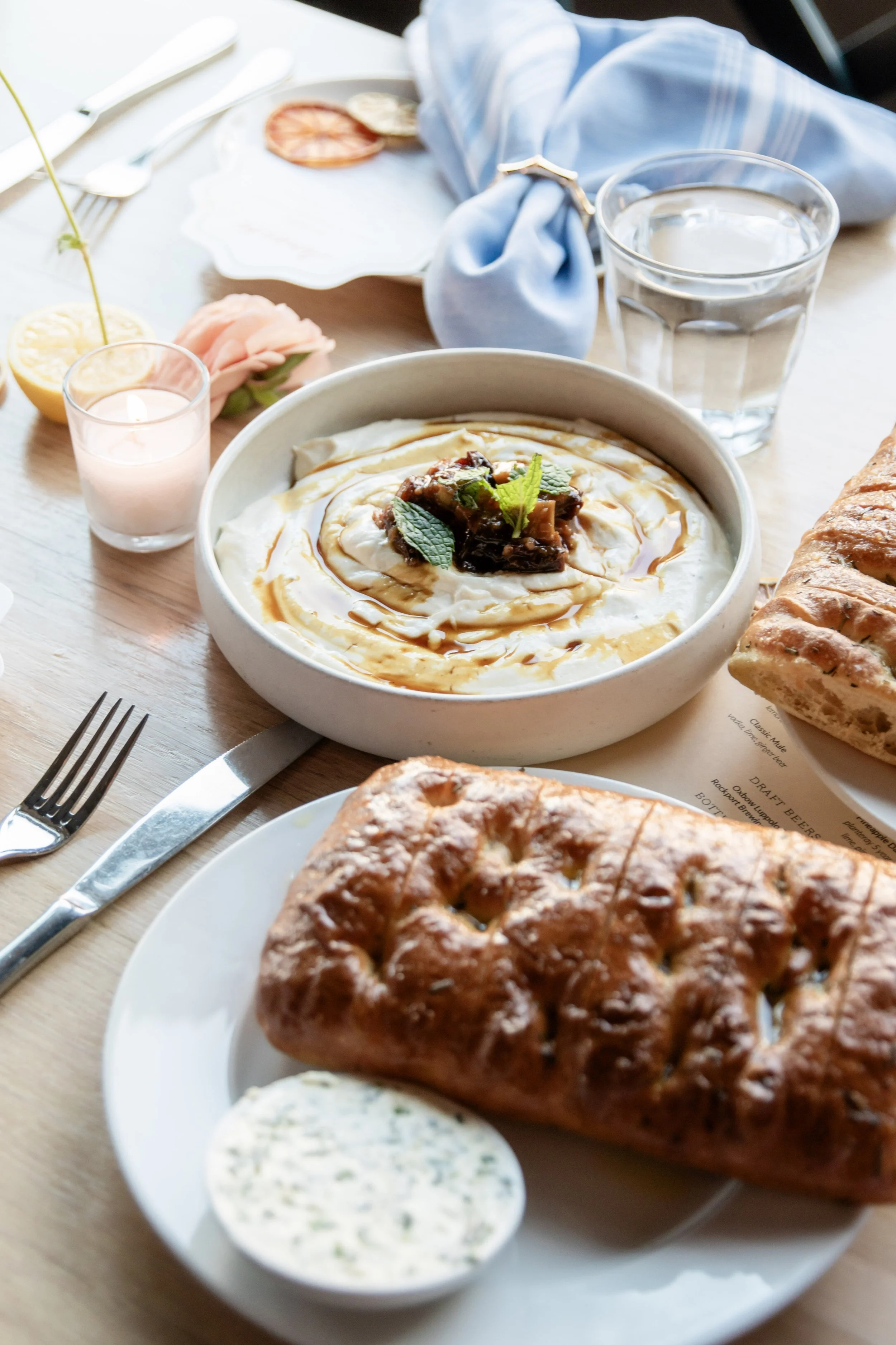 Close-up of a breakfast table with a plate of creamy yogurt topped with fruit and syrup, a loaf of bread, a small dish of herb butter, a glass of water, a candle, and some decorative flowers, set on a wooden table.