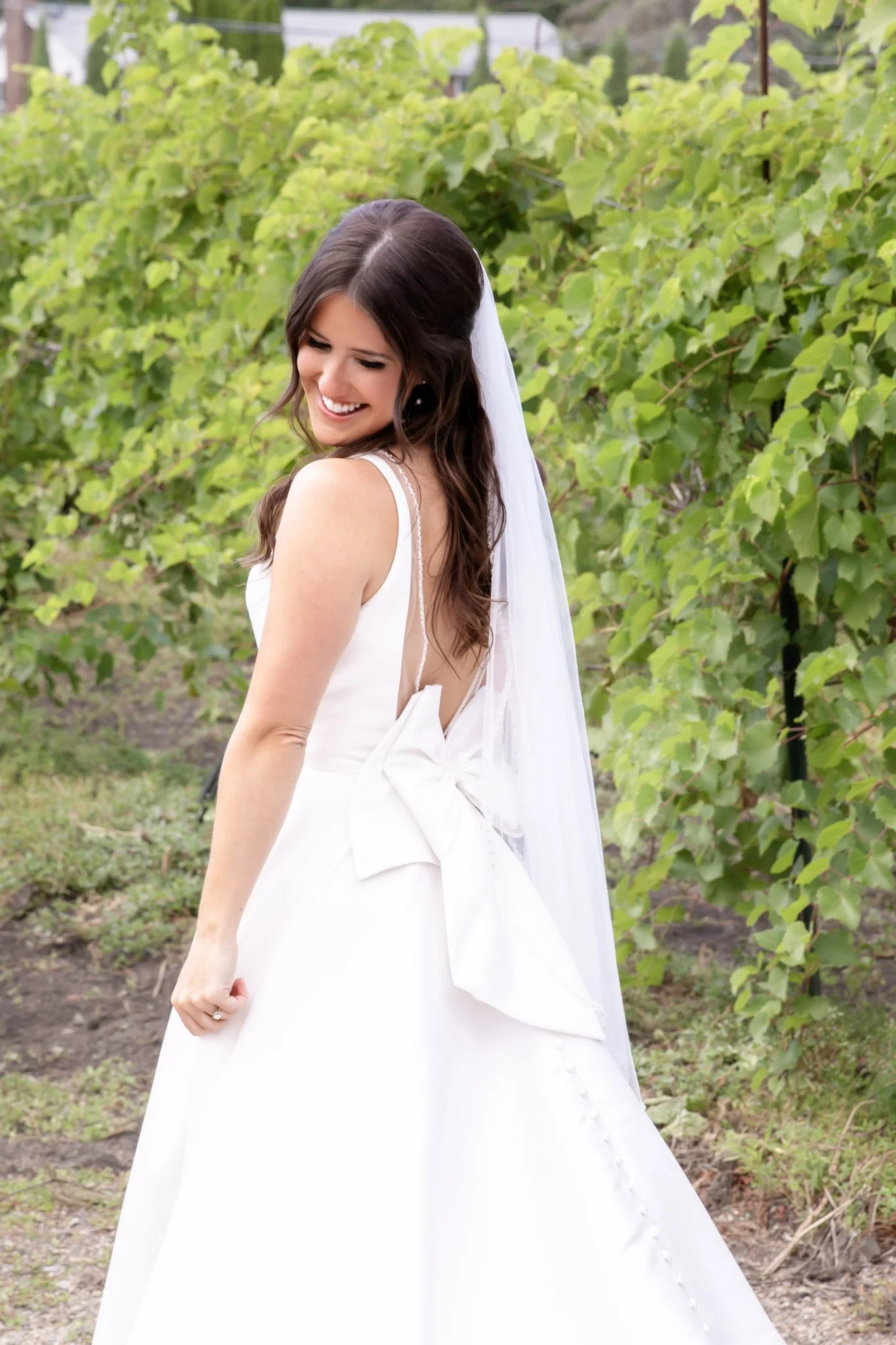 A smiling bride in a white wedding dress with a large bow detail on the back, standing outdoors among green bushes.