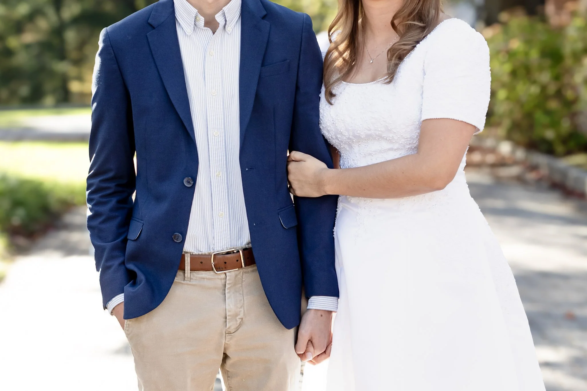 A couple dressed in formal attire standing outdoors, with the man's arm around the woman's waist. The man is wearing a blue blazer, striped shirt, and beige pants, while the woman is in a white wedding dress with short sleeves.