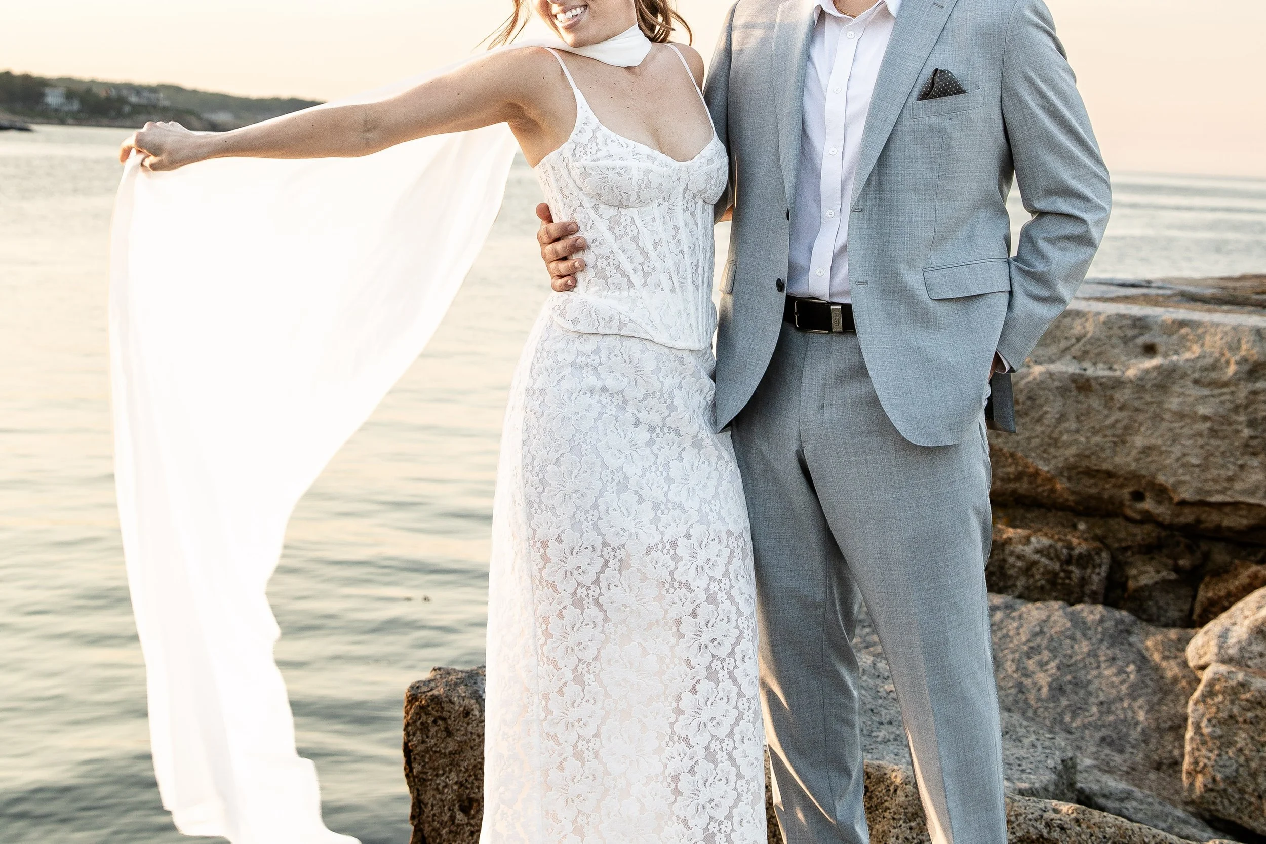 A couple standing together by the water during sunset, with the woman in a white lace dress and the man in a gray suit.