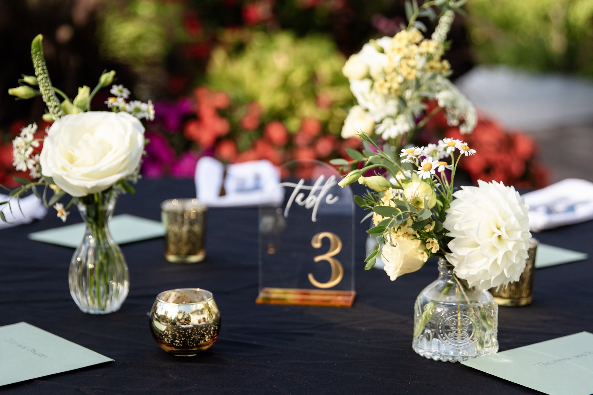 Table decorated with vases of white flowers, gold candle holders, and a table number sign at an outdoor event.