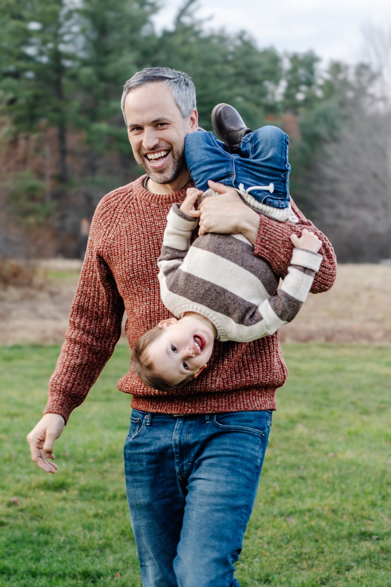 A man smiling and carrying two children, one upside down, in an outdoor field with trees in the background.