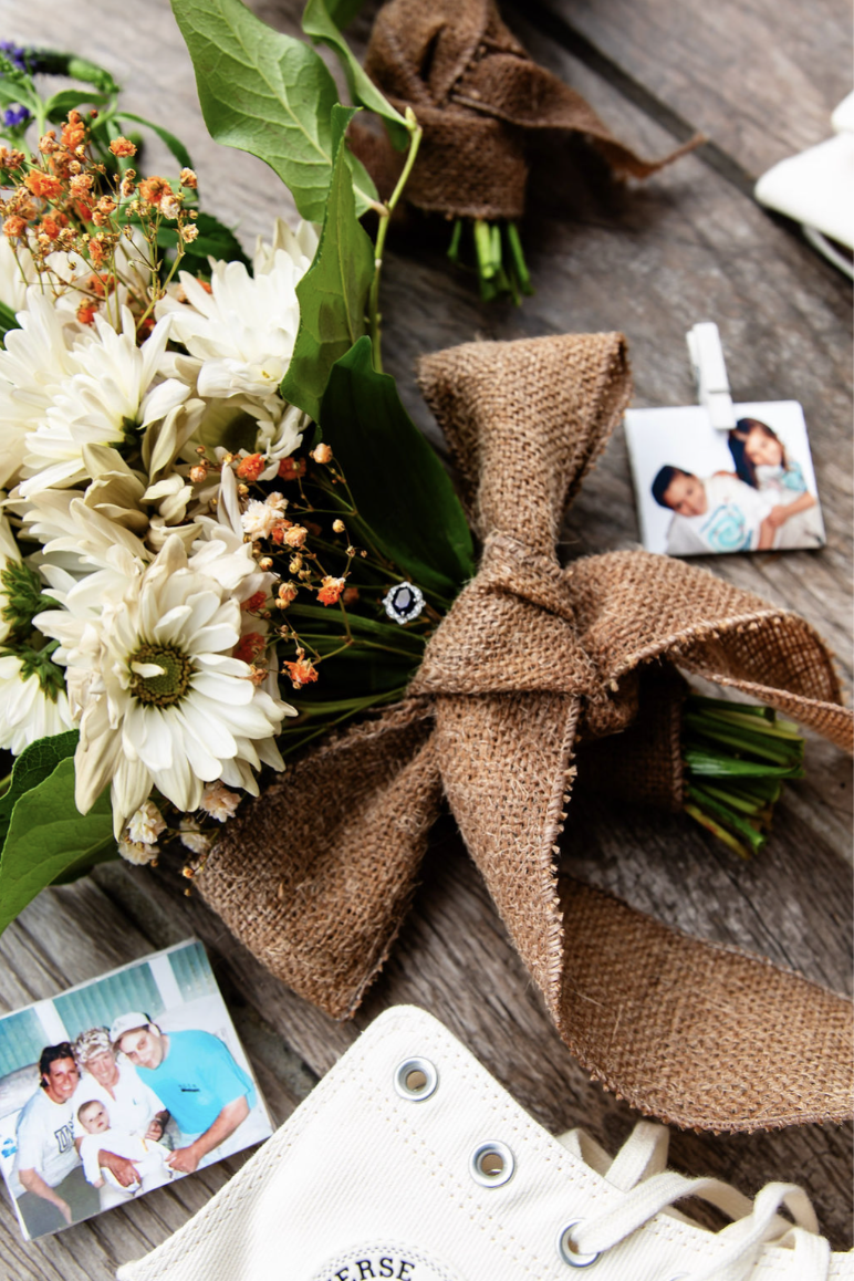 A bouquet of white daisies and other flowers with a brown burlap bow on a wooden surface, surrounded by small photographs and a pair of white Converse sneakers.