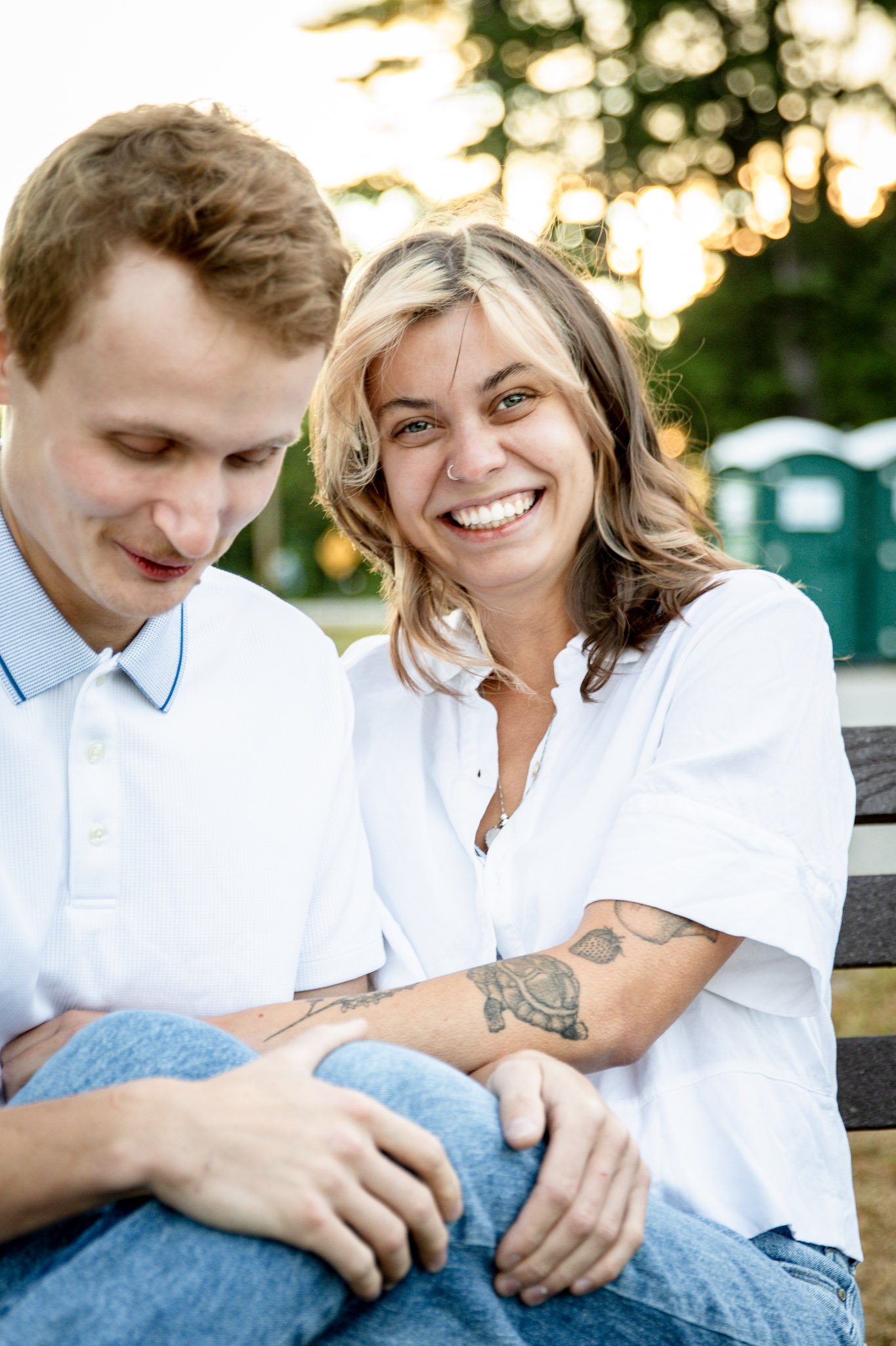 Two smiling women sitting on a park bench, one with tattoos, holding hands and looking down, the other smiling at the camera, during sunset.