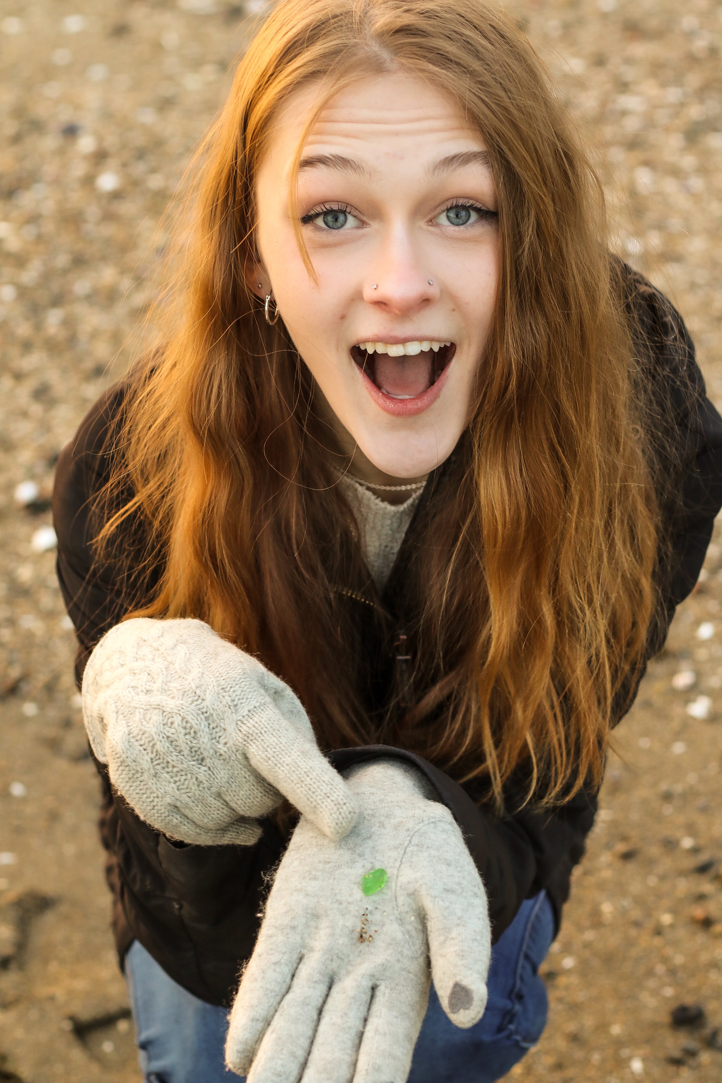 A young woman with long red hair and fair skin, wearing a beige glove, black jacket, and earrings, is outdoors on a dirt ground, holding a small green sea glass piece in her gloved hand, with a surprised and excited expression on her face.