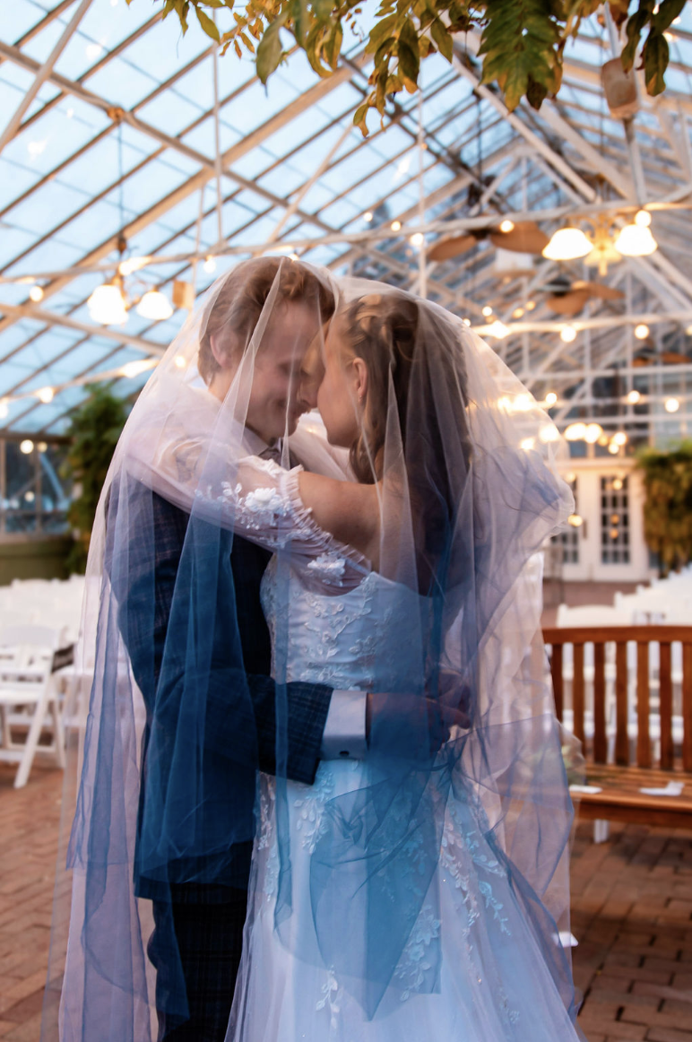 A bride and groom share an intimate moment under a veil at their wedding reception inside a greenhouse or glass conservatory with string lights and greenery.