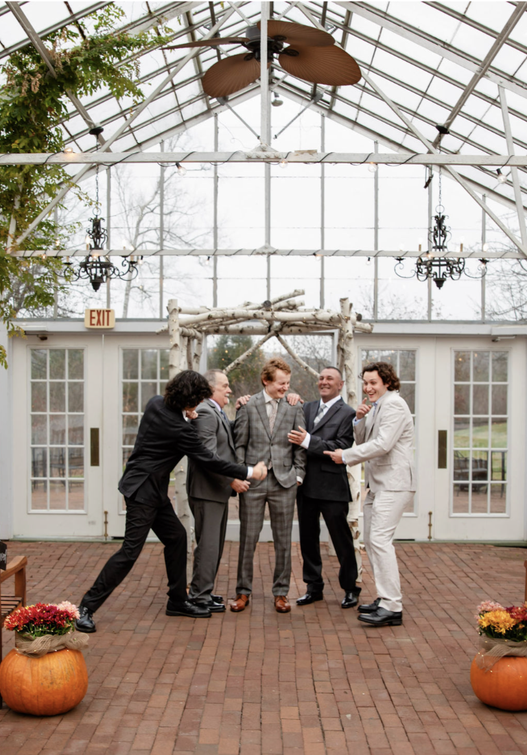 Six men dressed in suits, laughing and engaging in playful interaction inside a greenhouse with large windows and brick flooring, decorated with pumpkins and flowers.
