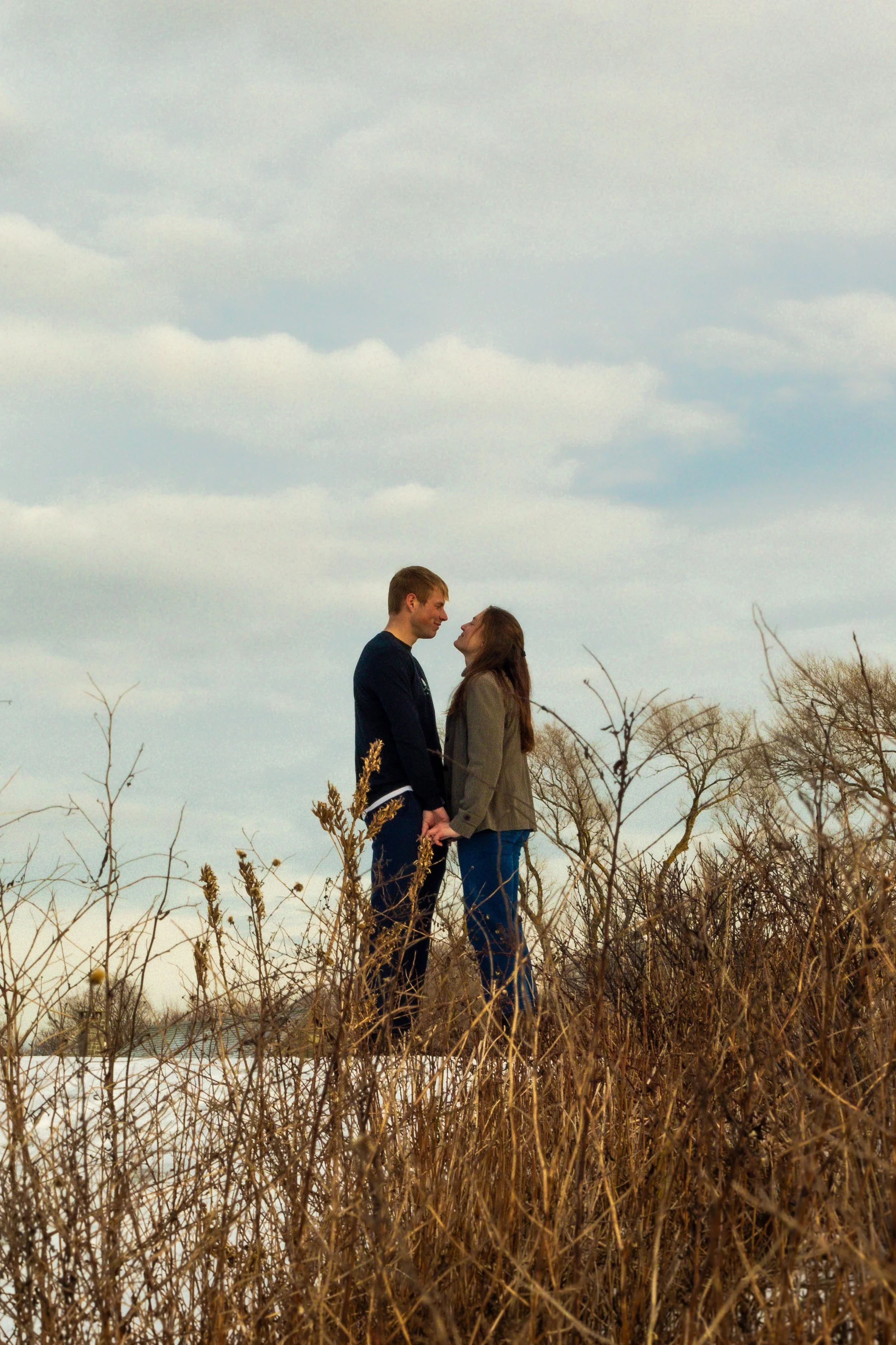 A couple standing close together in a field with dry grass, holding hands and gazing into each other's eyes under a cloudy sky.
