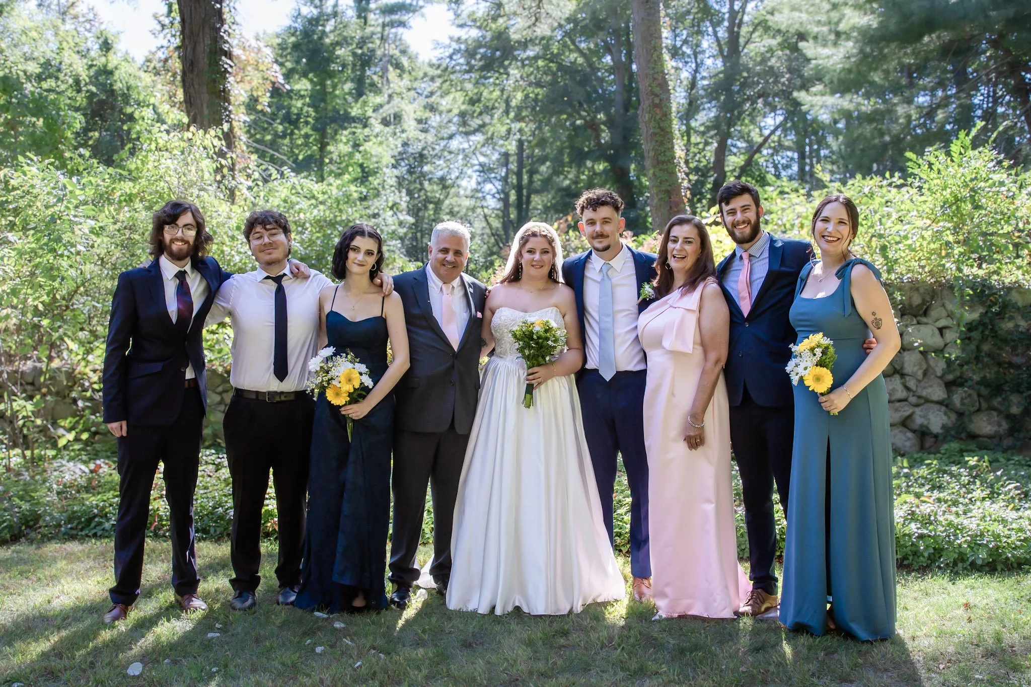 Group of wedding guests posing together outdoors on a sunny day, surrounded by greenery and trees. The group includes men and women dressed in formal attire, with some holding bouquets of flowers.