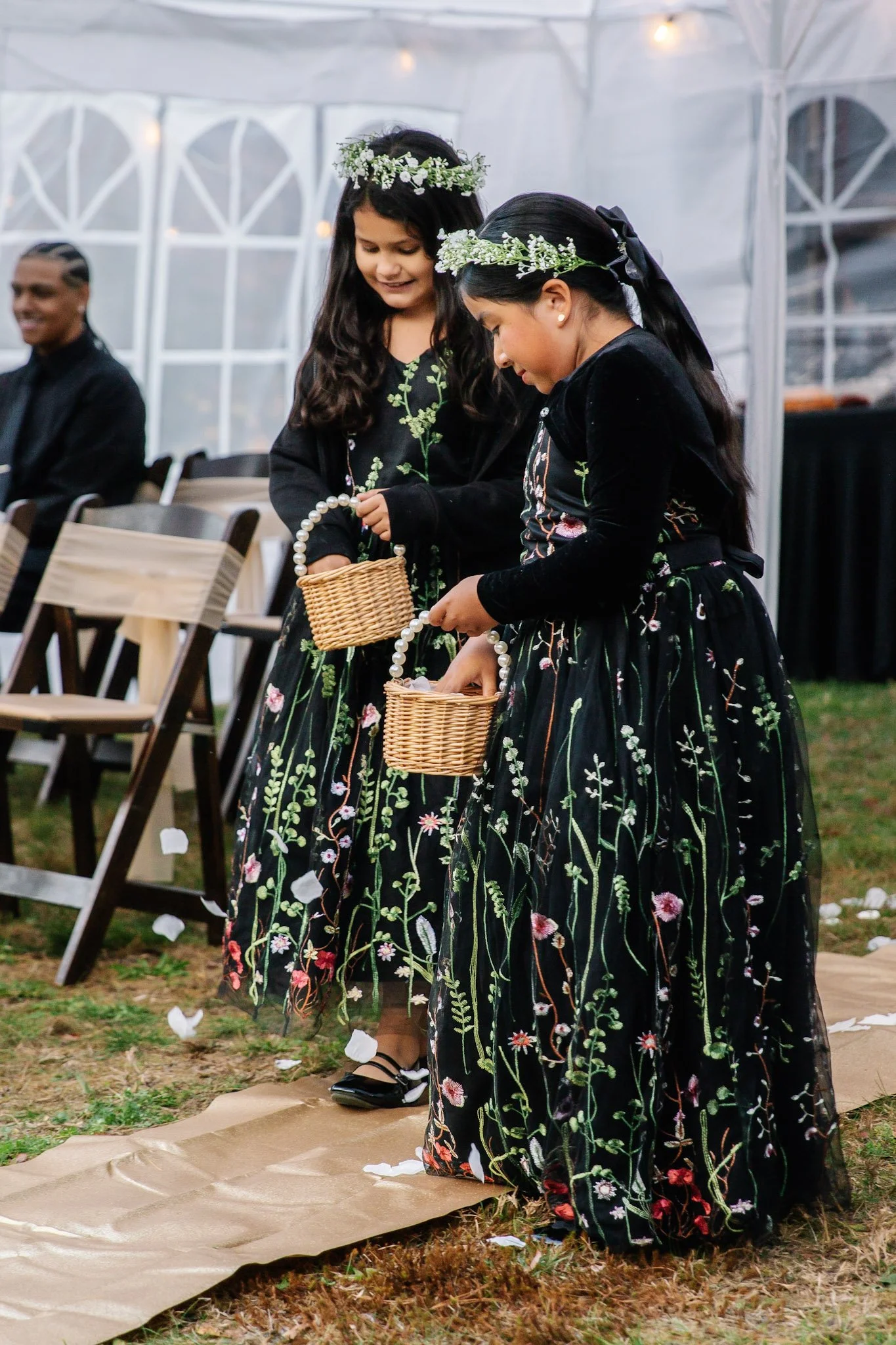 Two young girls dressed in black floral dresses with flower crowns, holding small wicker baskets, participating in a wedding ceremony outdoors.