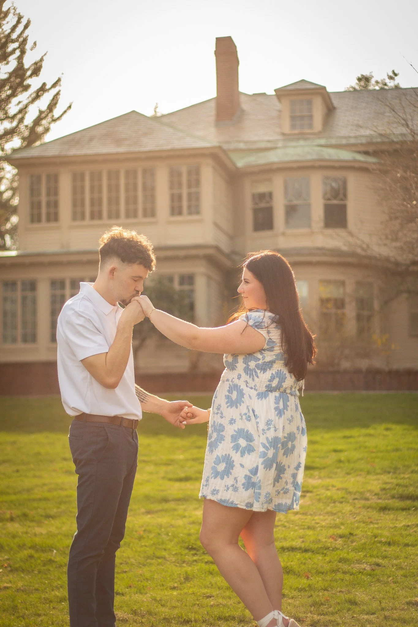A couple standing on a lawn is engaged in a romantic moment, with the man kissing the woman's hand and the woman smiling at him, in front of a large house.
