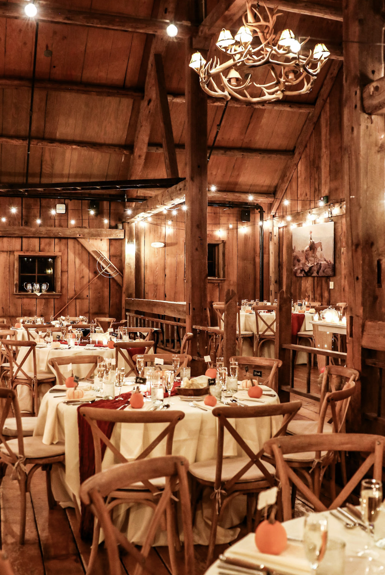 Decorated rustic wooden event space with round tables covered in cream tablecloths, set with glasses and orange pumpkins, string lights, and a chandelier with antlers hanging from a wooden ceiling.