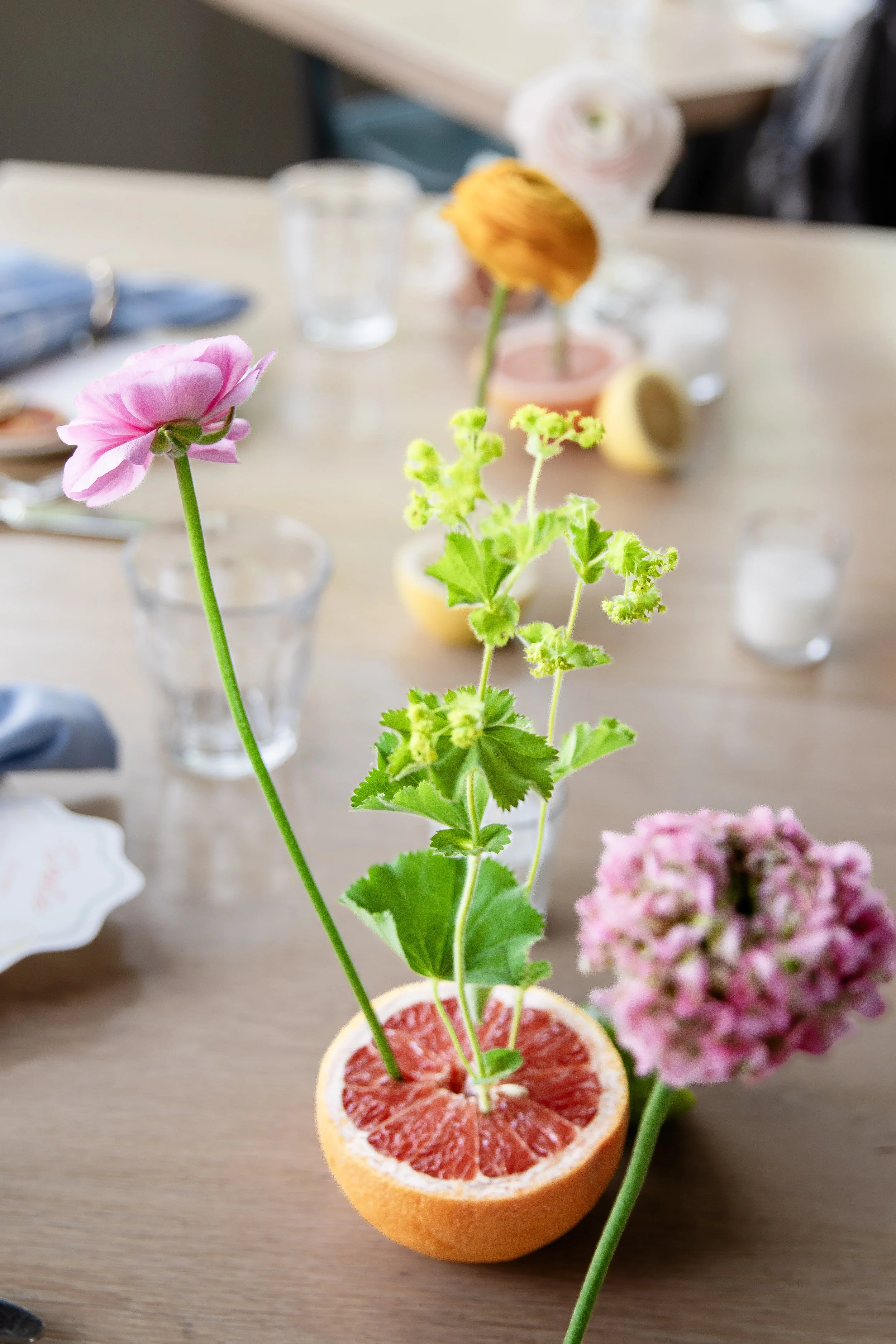 A small indoor plant in a container filled with red soil, with pink and yellow flowers, set on a wooden table. In the background, there are various other flowers, glasses, and objects on the table.