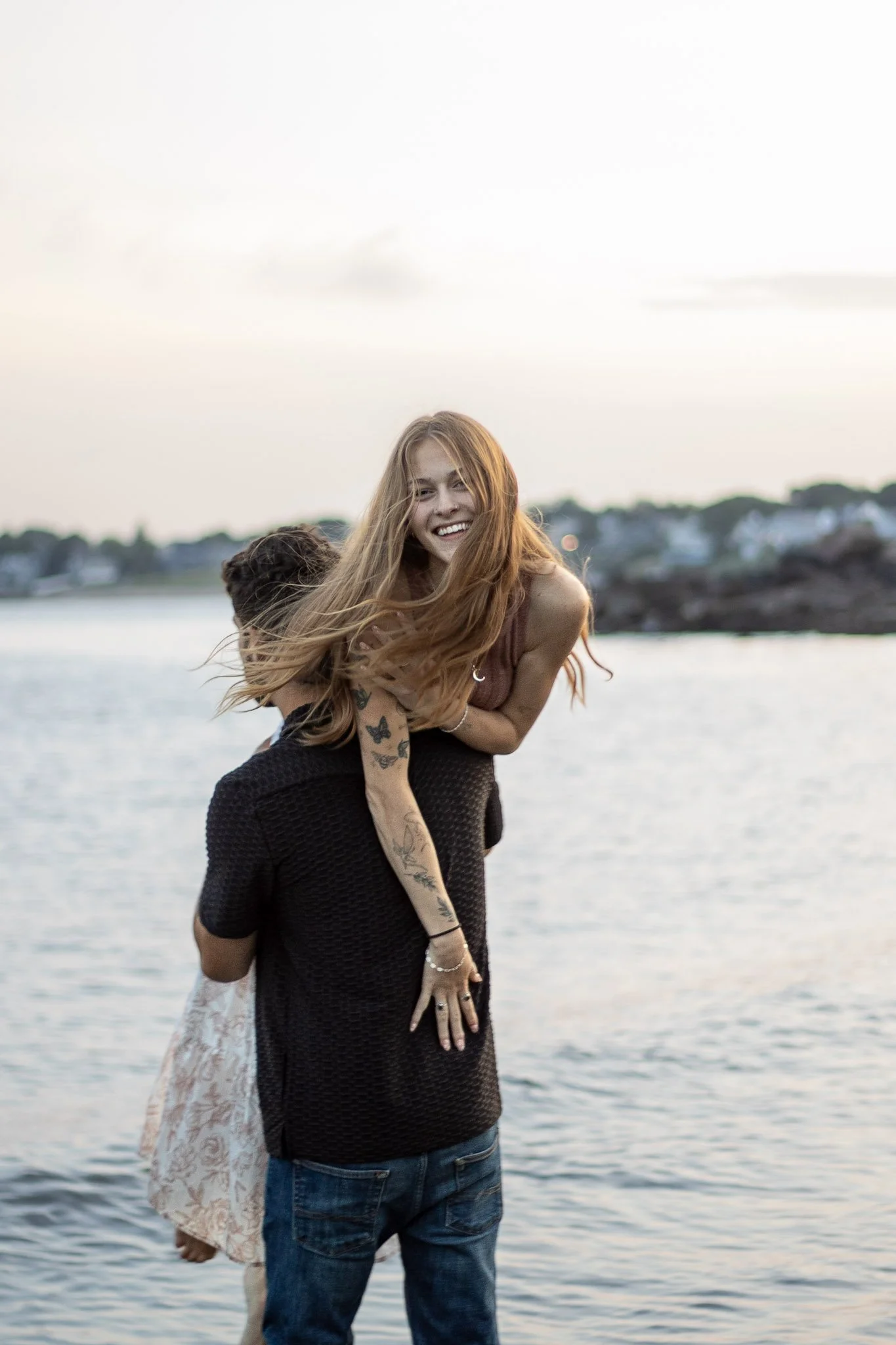 A man is carrying a woman on his shoulders by the water, both are smiling and having fun during sunset.