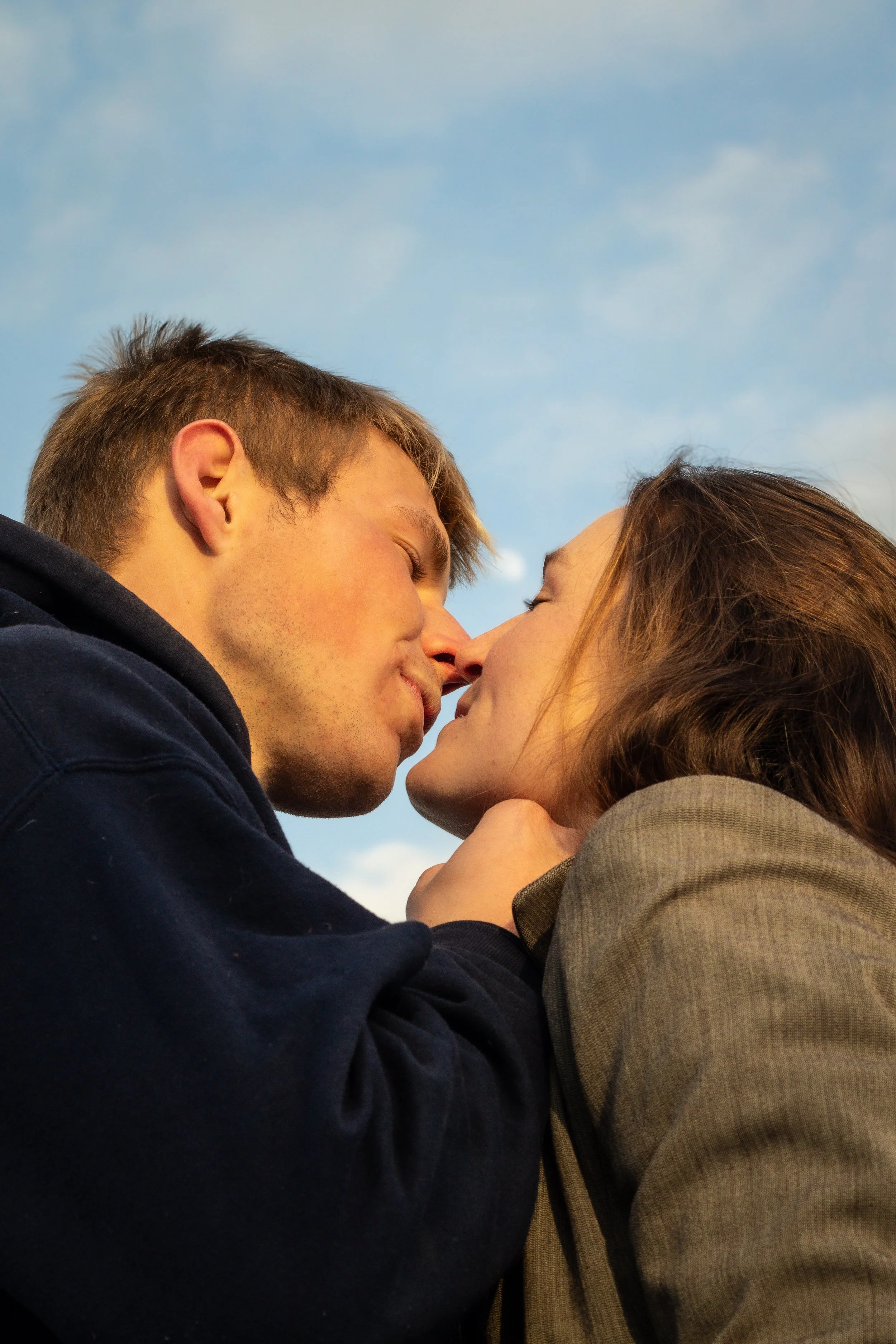 A young couple sharing a kiss outdoors under a blue sky.