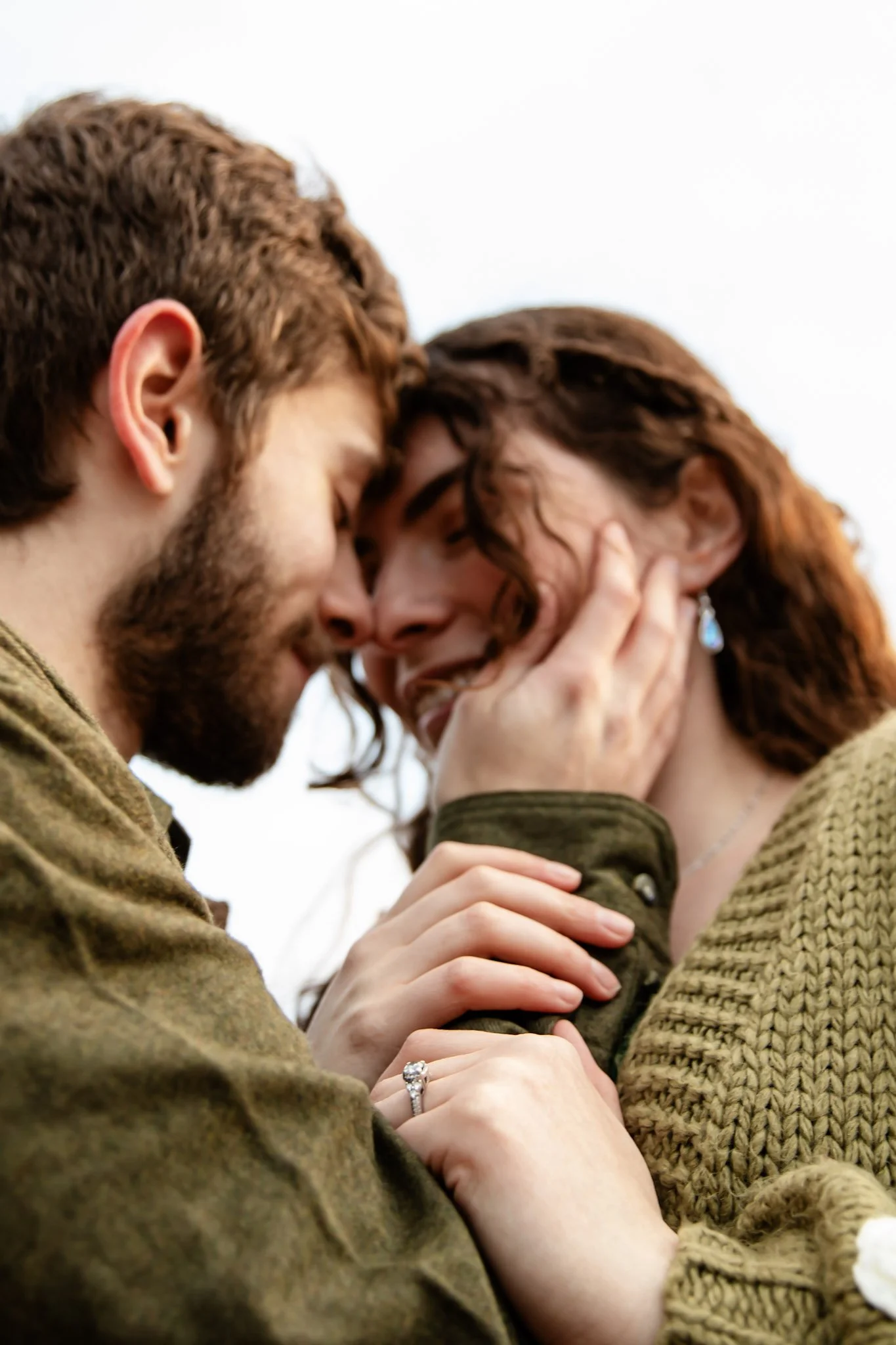 Close-up of a young couple with their foreheads touching, smiling, and holding hands, with a ring on the woman's finger.