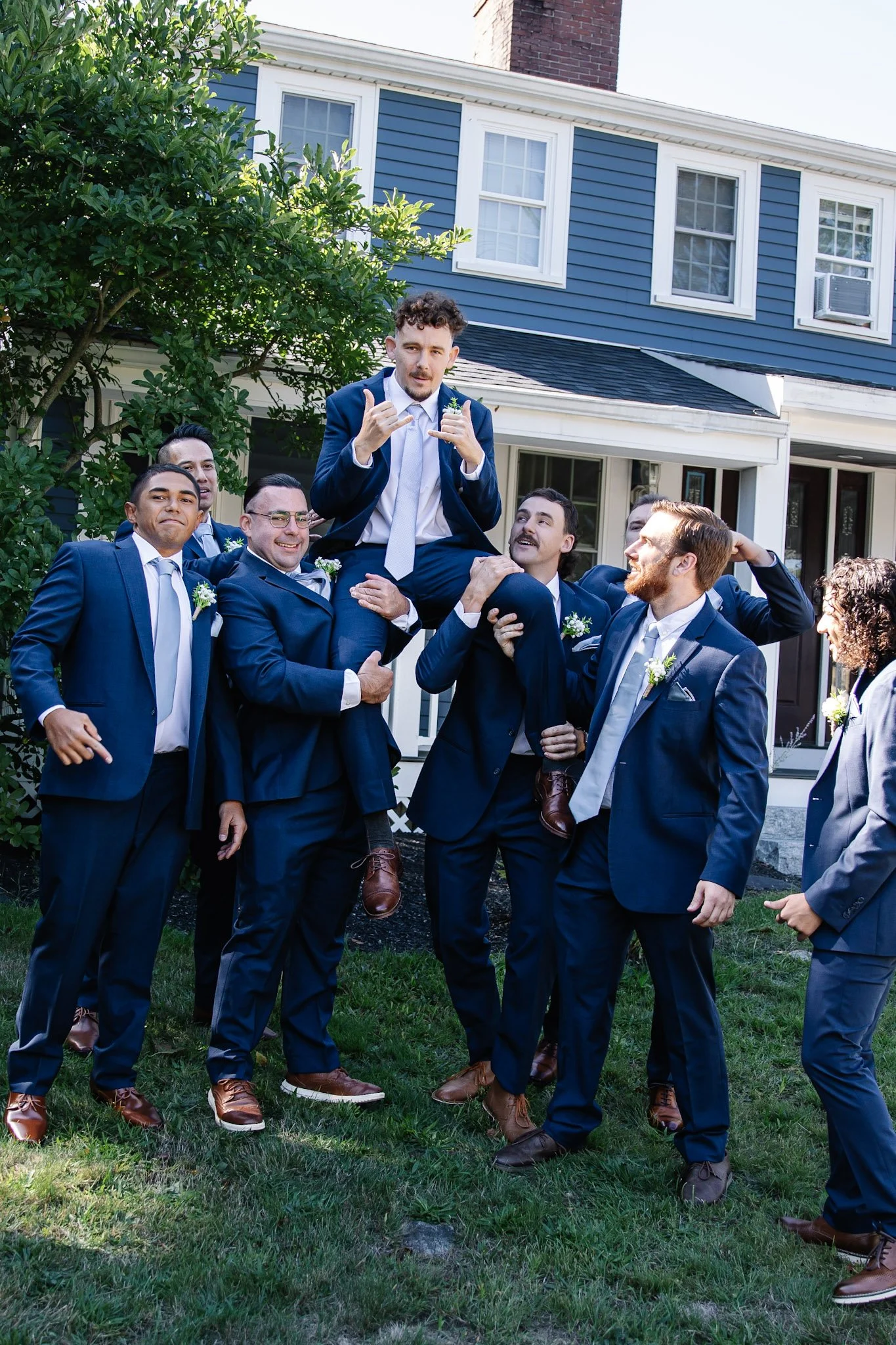 A group of men in navy suits celebrating outdoors, with one man being lifted on others' shoulders during a wedding or special event.