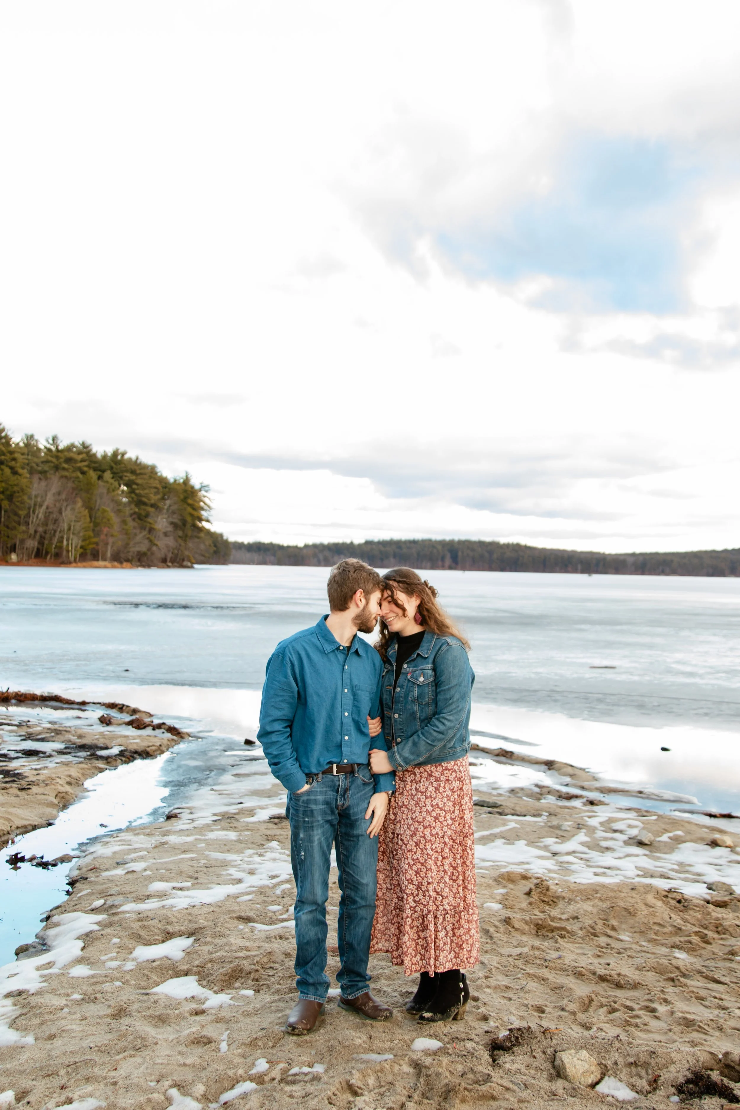 A couple standing close together on a sandy beach near a frozen lake, sharing an intimate moment with their foreheads touching, surrounded by a natural landscape of trees and cloudy sky.