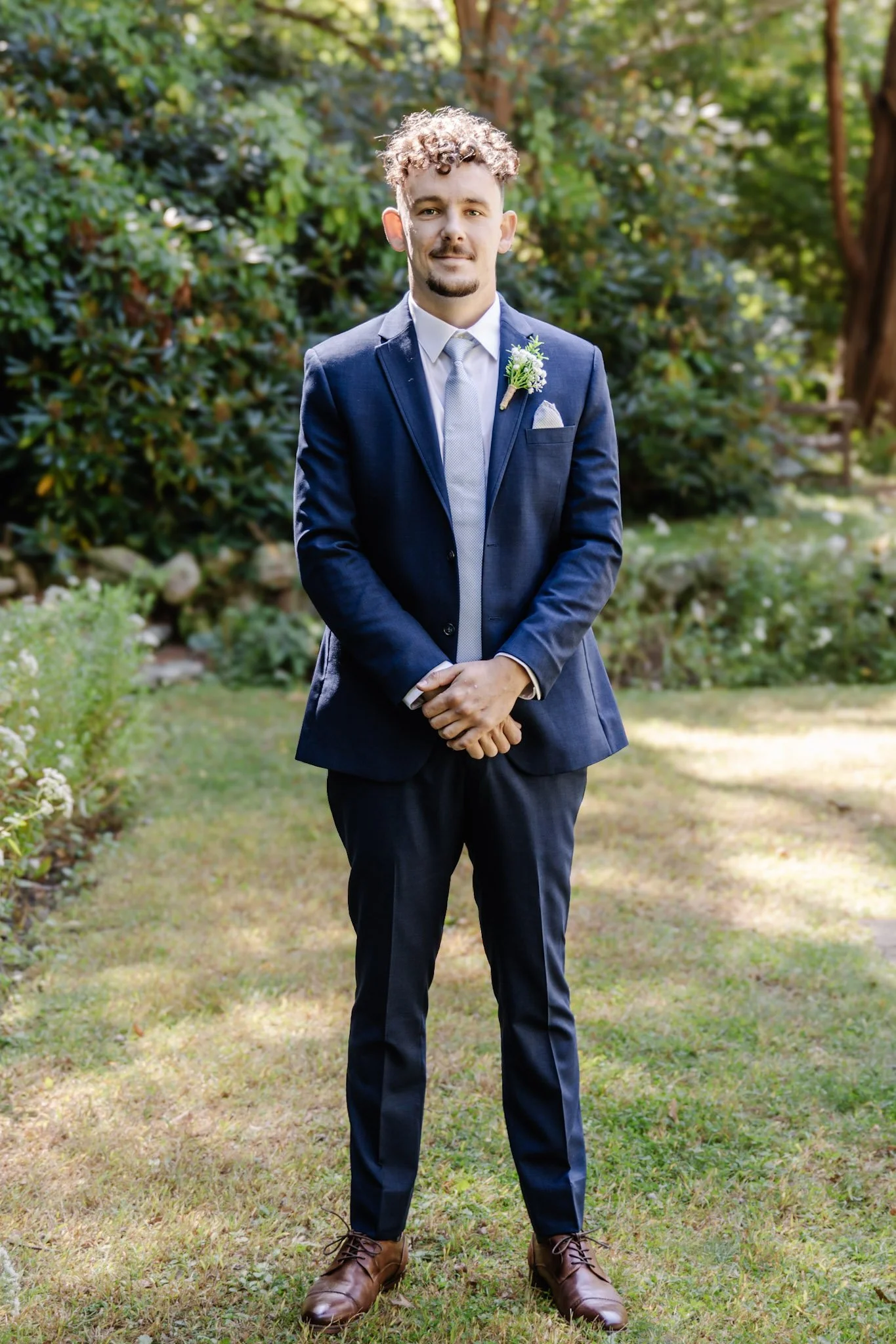 A man in a navy suit and tie standing outdoors on grass with green trees and bushes in the background, dressed formally with a boutonniere on his lapel.
