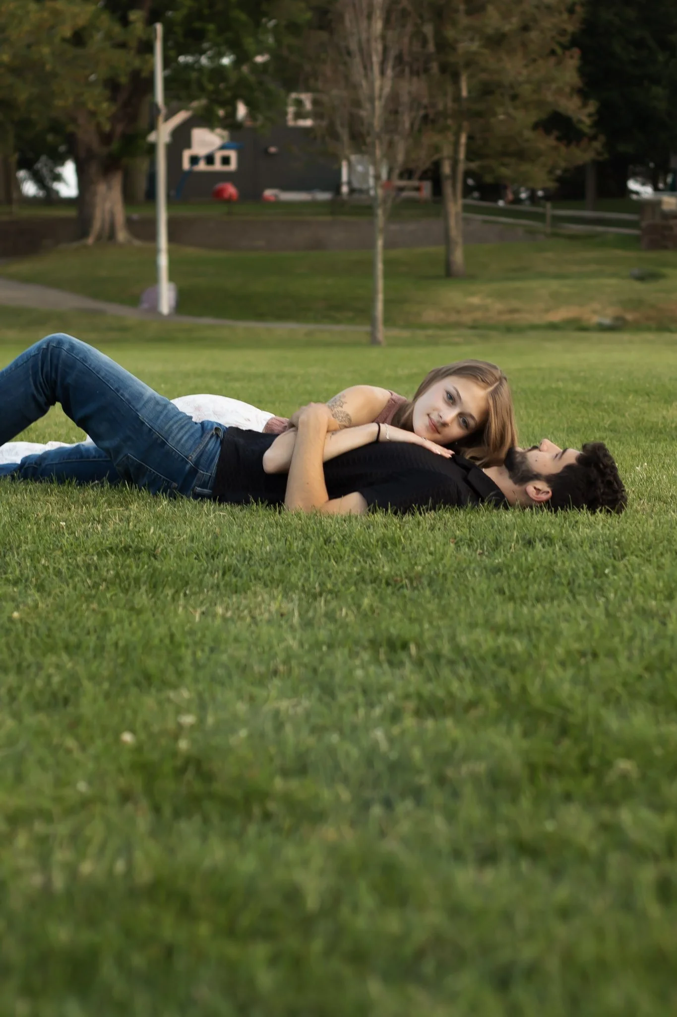 A young woman with blonde hair lying on top of a young man with a beard on a grassy field, looking affectionate. The man is lying on his back, and the woman is resting on him, with her head on his chest. Trees and houses are visible in the background
