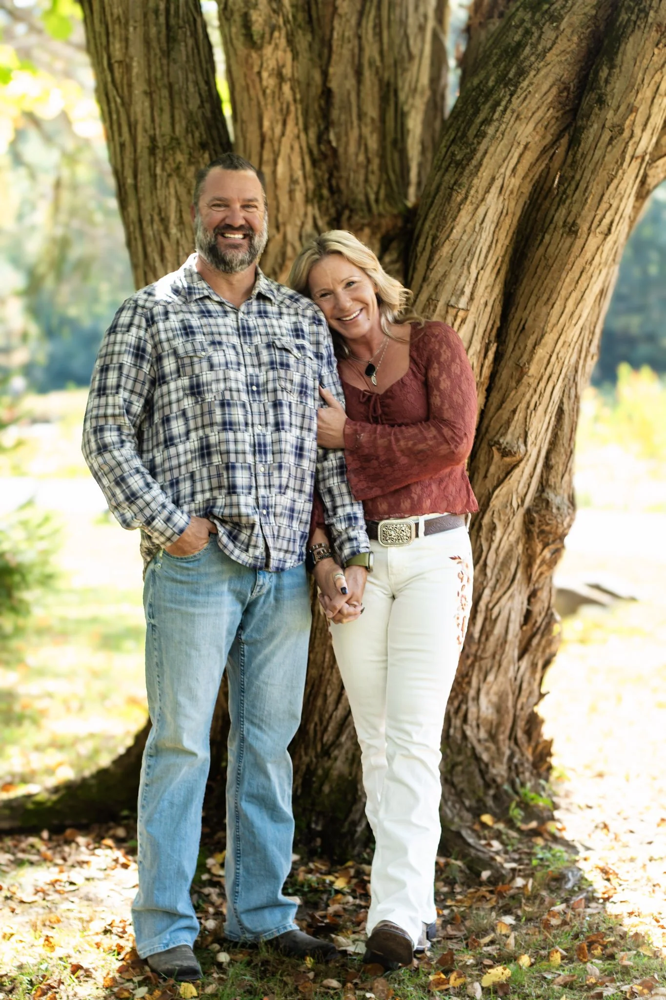 A smiling man and woman standing close together outdoors near a large tree, holding hands and leaning into each other, surrounded by fallen autumn leaves.