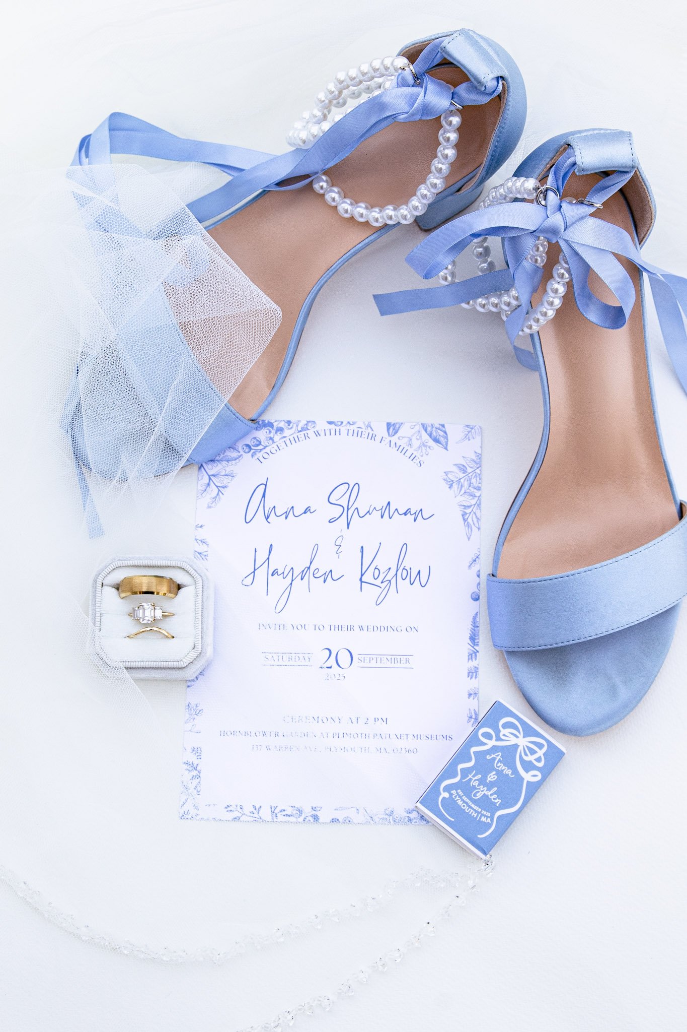Cape Cod, MA Luxury Wedding Invitation flatlay. Light blue high heel shoes with pearl ankle straps and ribbons, a wedding invitation, gold wedding rings in a ring box, and a ring with a large stone.