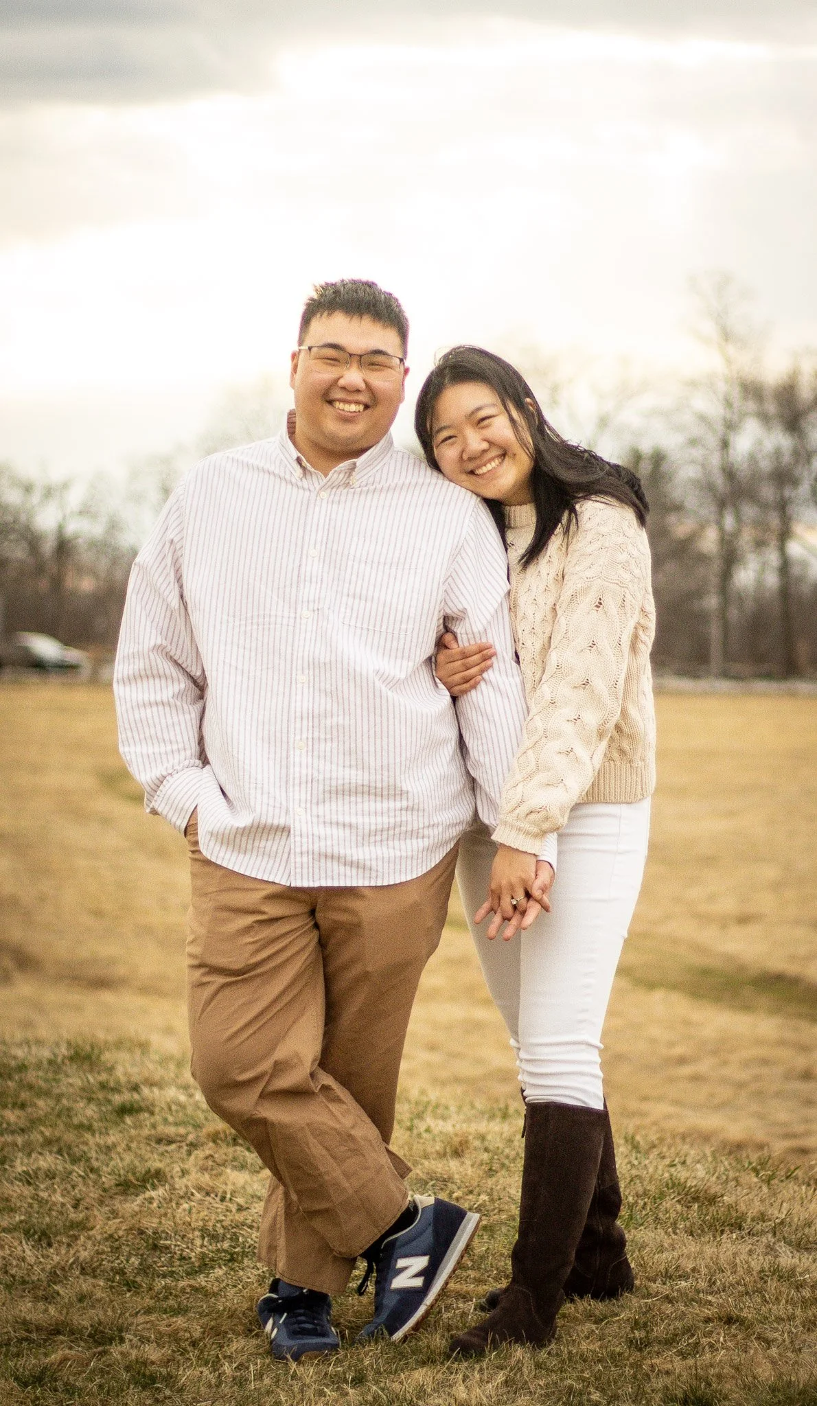 A smiling couple holding hands in a grassy outdoor field with trees and a cloudy sky in the background
