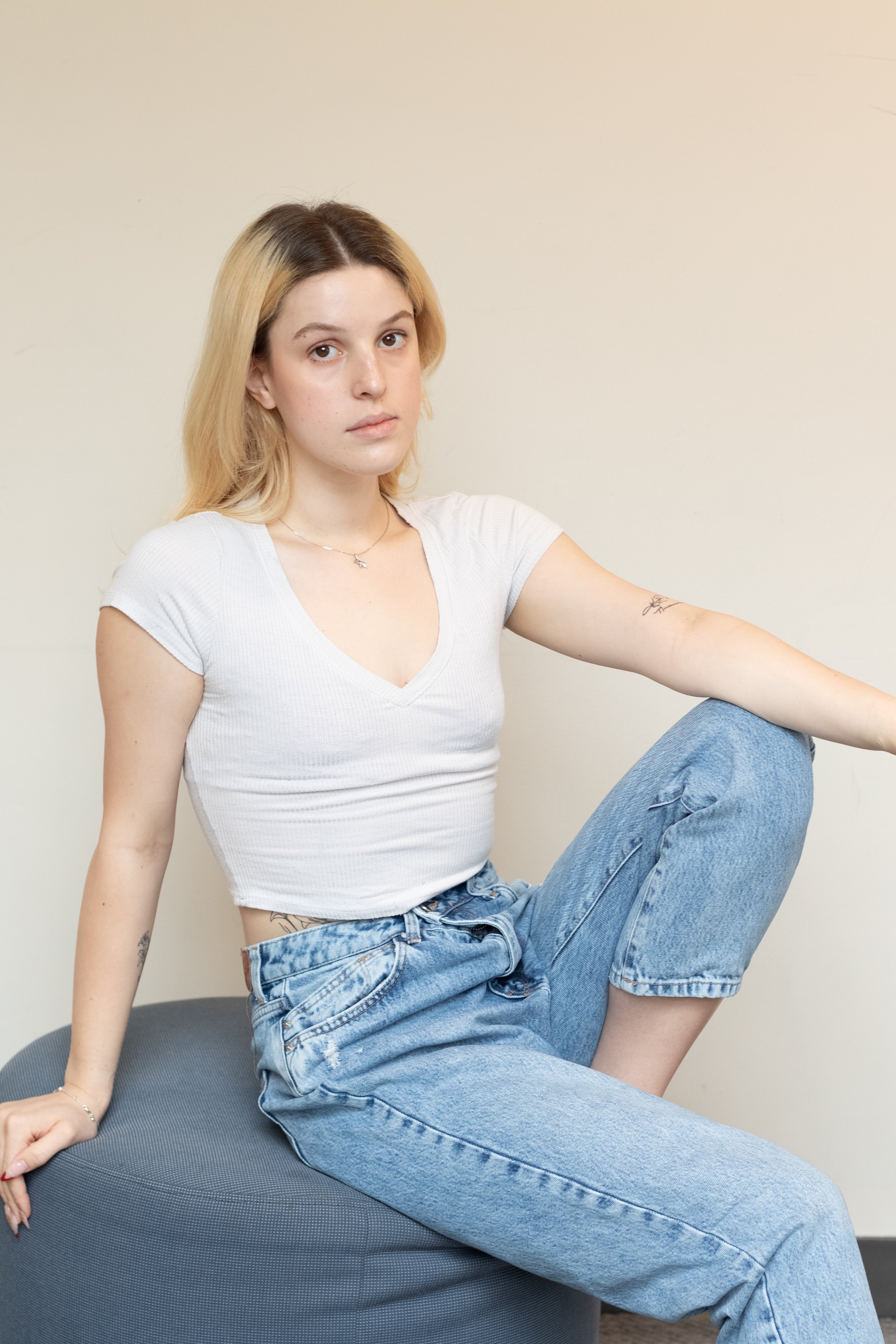 Boston, MA, Young woman with blonde hair, wearing a white t-shirt and blue jeans, sitting on a black cushion against a plain beige wall, looking directly at the camera.