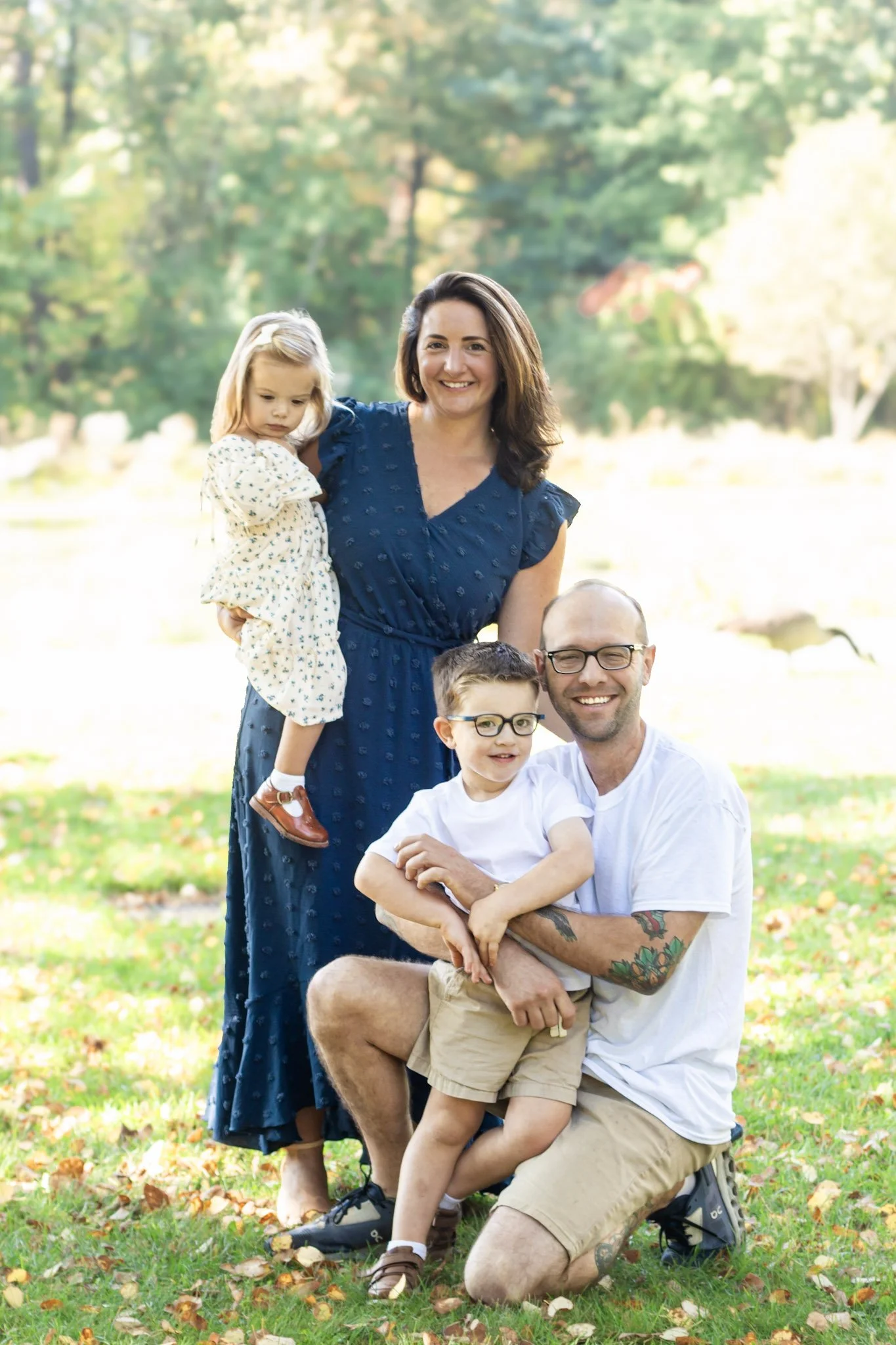 Southern NH, Greater Manchester, NH, A smiling family group outdoors in a park with green trees and fallen leaves on the ground. The group consists of a woman, a man, and three children, one of whom is being held by the woman.