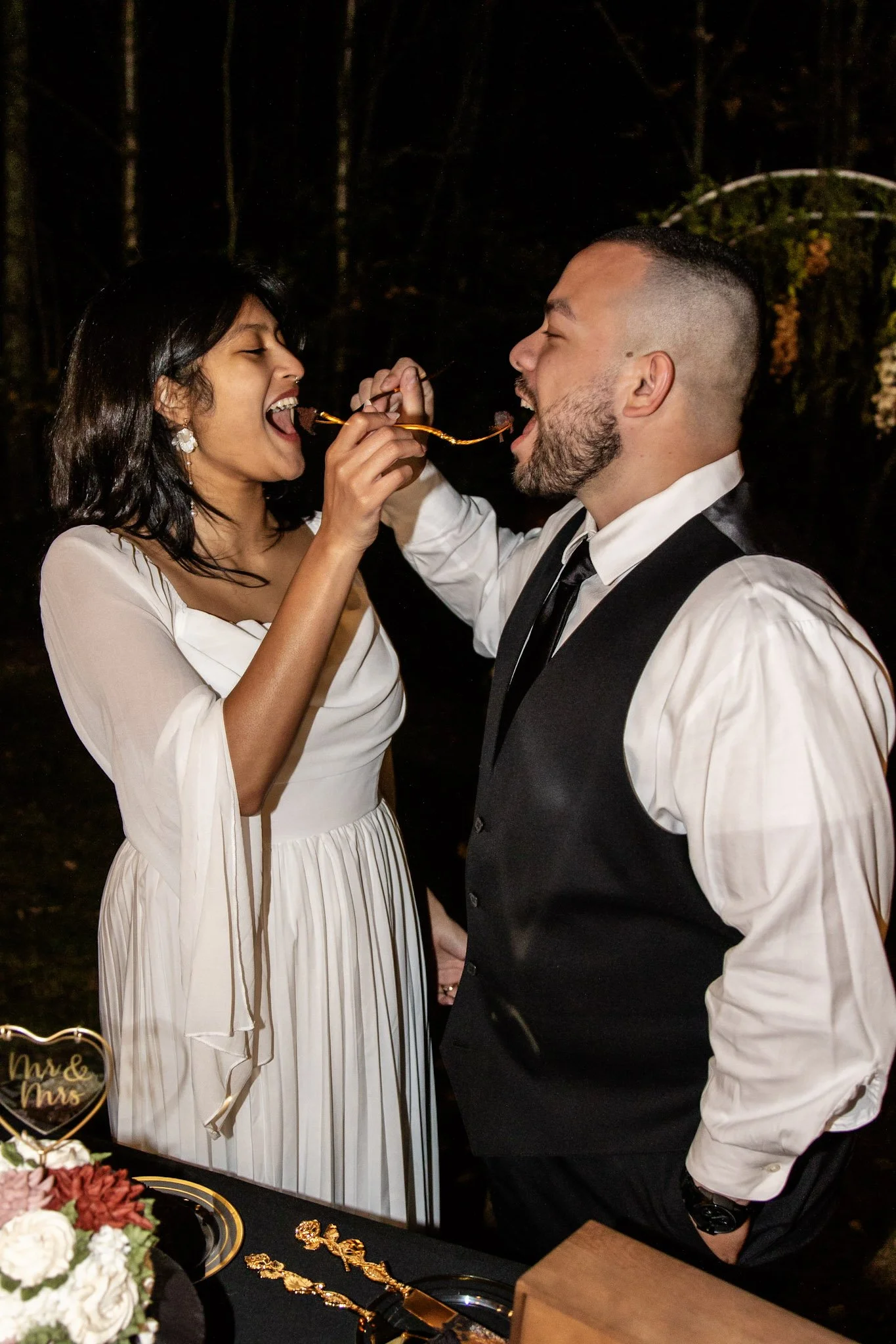 A couple sharing a piece of cake at a wedding reception, with the bride in a white dress and the groom in a white shirt with a black vest, in a dimly lit outdoor setting.