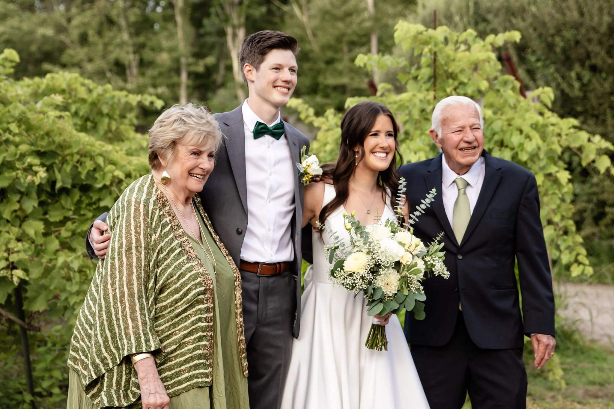 Group of four people smiling outdoors at a wedding, with a bride holding a bouquet, a groom wearing a suit and bow tie, and two elderly individuals, all standing in front of green foliage.