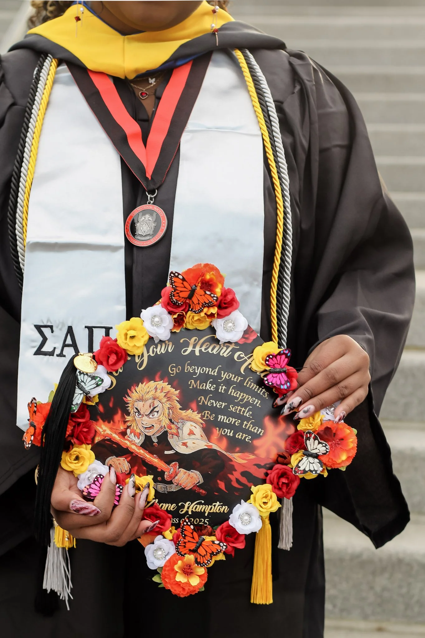 A person dressed in graduation regalia holding a decorated graduation cap with anime and butterfly motifs and a motivational quote.