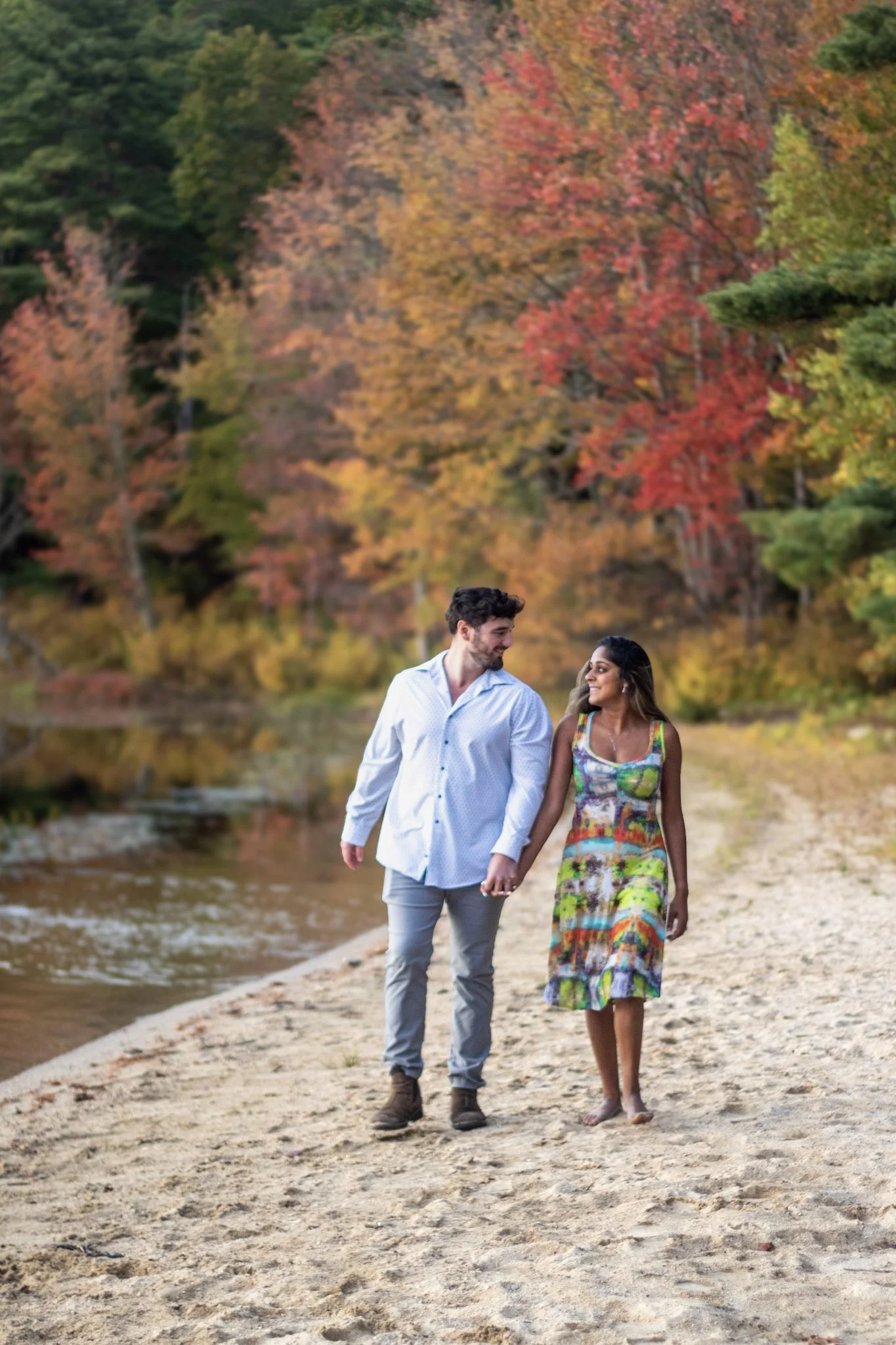 A couple walking hand in hand along a sandy riverbank, surrounded by colorful autumn trees.