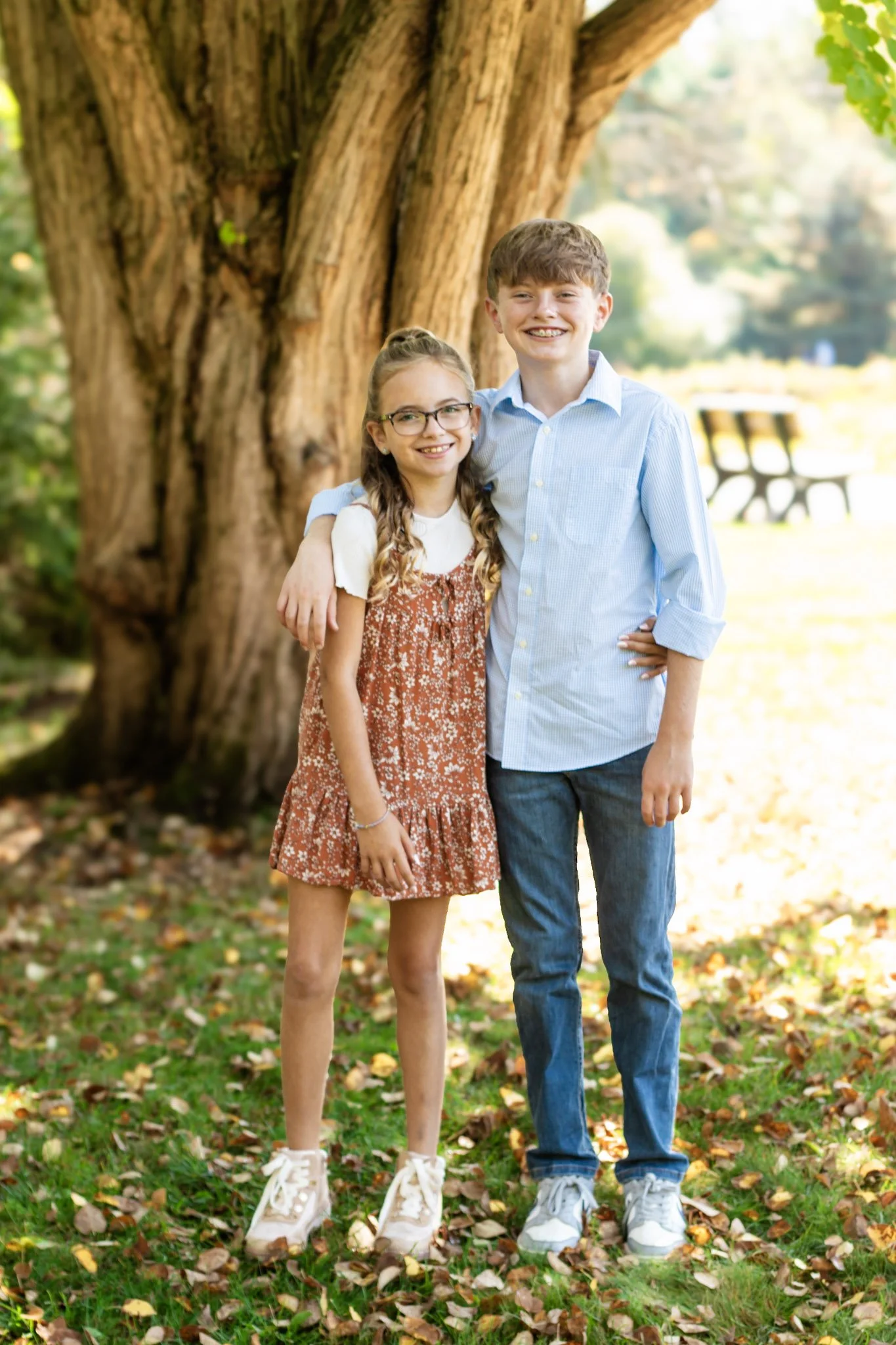 A young girl and boy standing outdoors near a large tree, smiling and posing for a photo. The girl has long hair in braids, glasses, and is wearing a white shirt with a rust-colored dress. The boy has curly hair, and is wearing a light blue dress shi