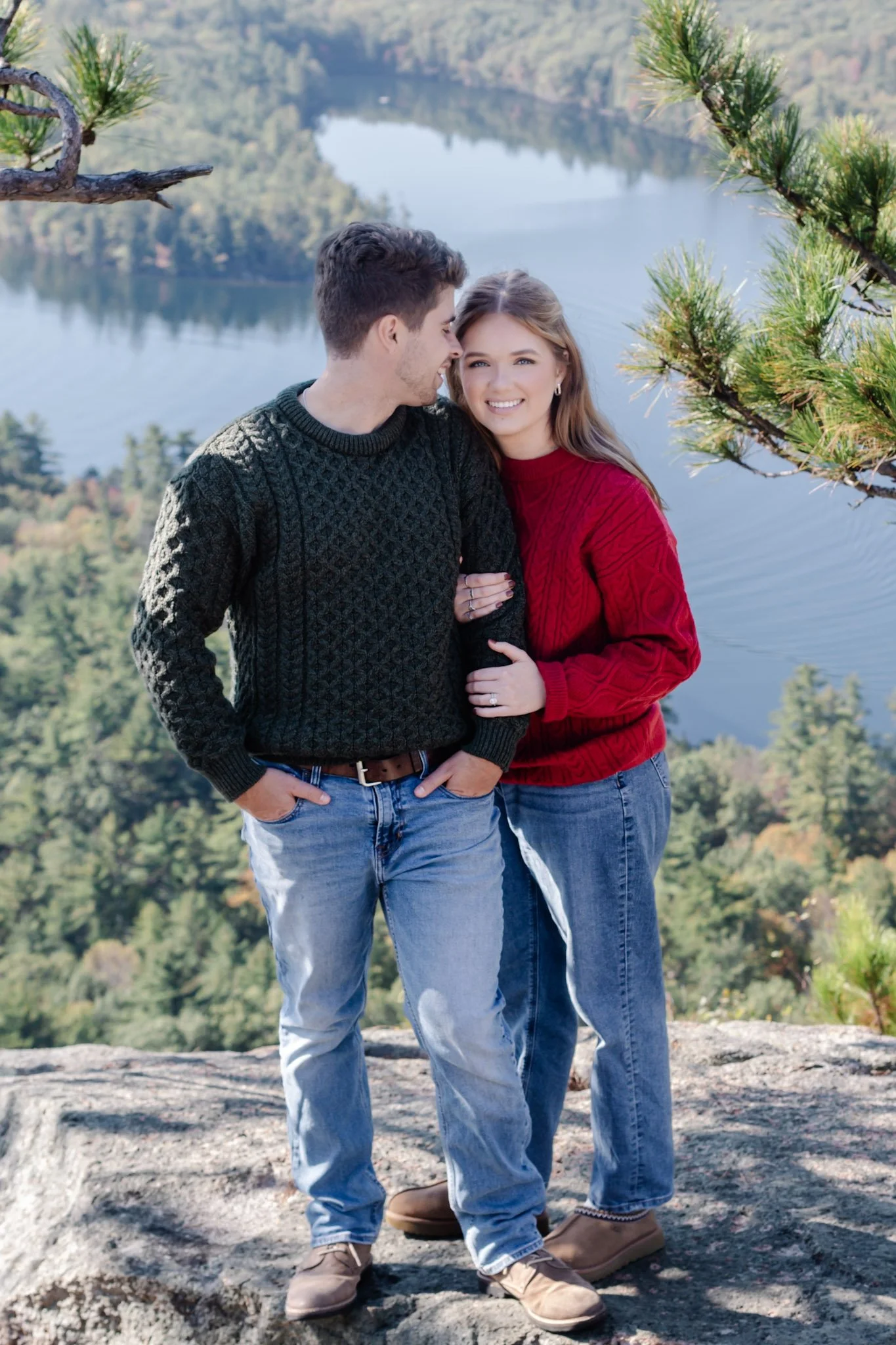 A couple stands close on a rocky outcrop with a scenic lake and forest in the background, smiling and leaning towards each other.