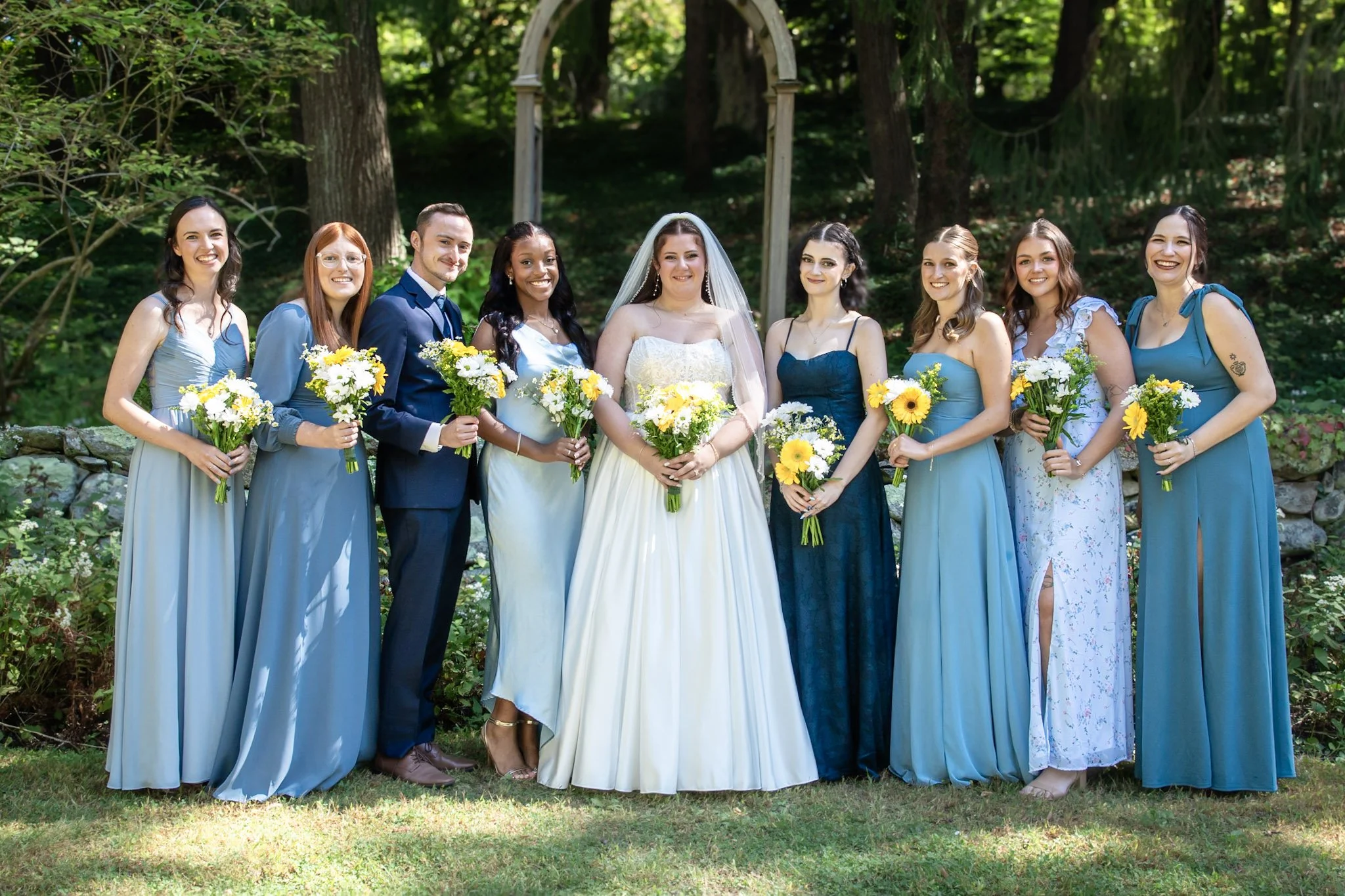 Bride in white wedding gown standing with her bridal party outdoors, surrounded by trees, all holding bouquets of yellow and white flowers.