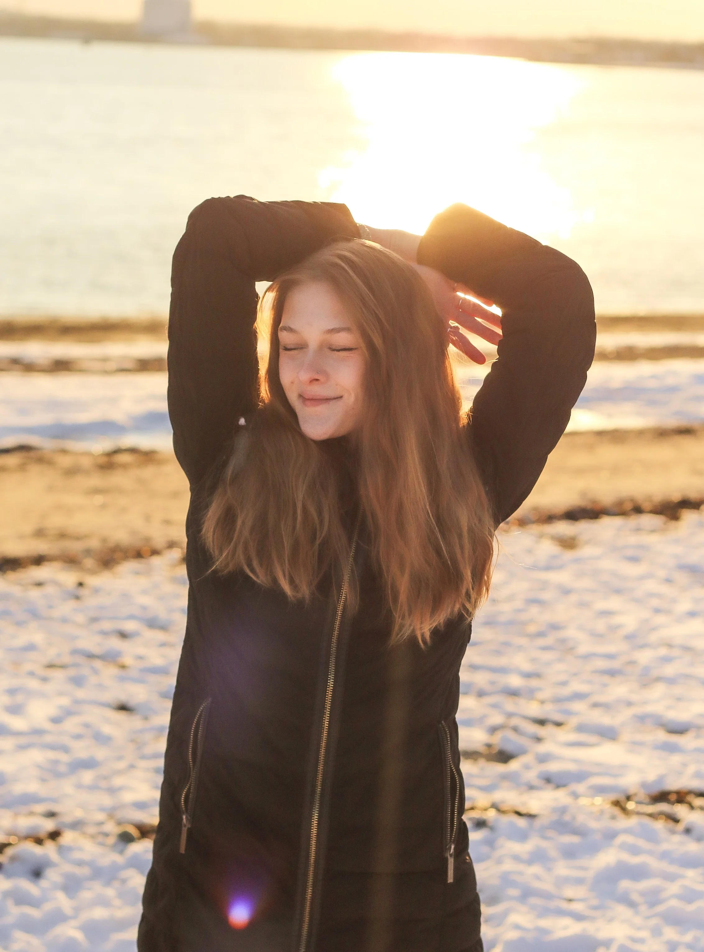 A young woman with long, wavy hair standing on a snowy beach during sunset, smiling with her eyes closed, wearing a black jacket, with the sun reflecting on the water behind her.