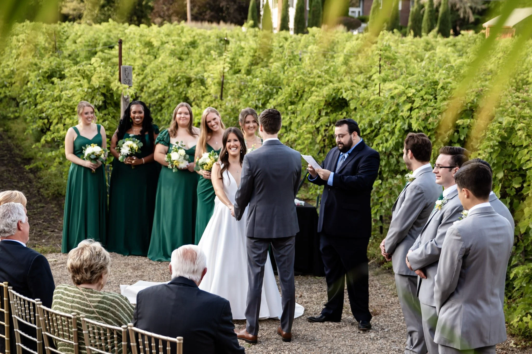 A fall vineyard wedding ceremony with a officiant reading from a paper, a bride and groom facing each other, and bridesmaids and groomsmen standing nearby, surrounded by green foliage in Nashua, NH.