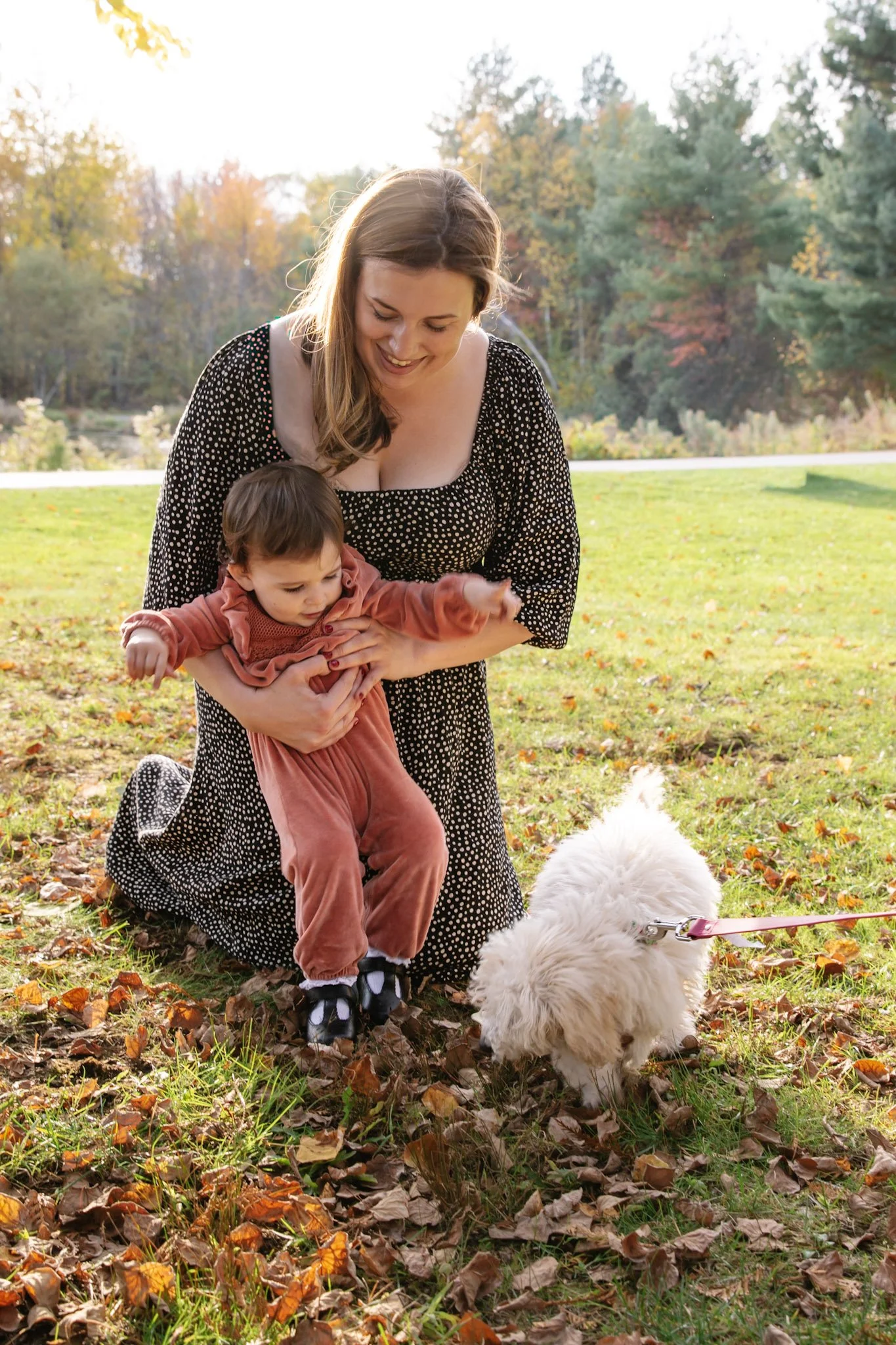 A woman and a young child are outside in a park with fall foliage. The woman is smiling and holding the child, who is reaching toward a small fluffy white dog on a leash sniffing fallen leaves.