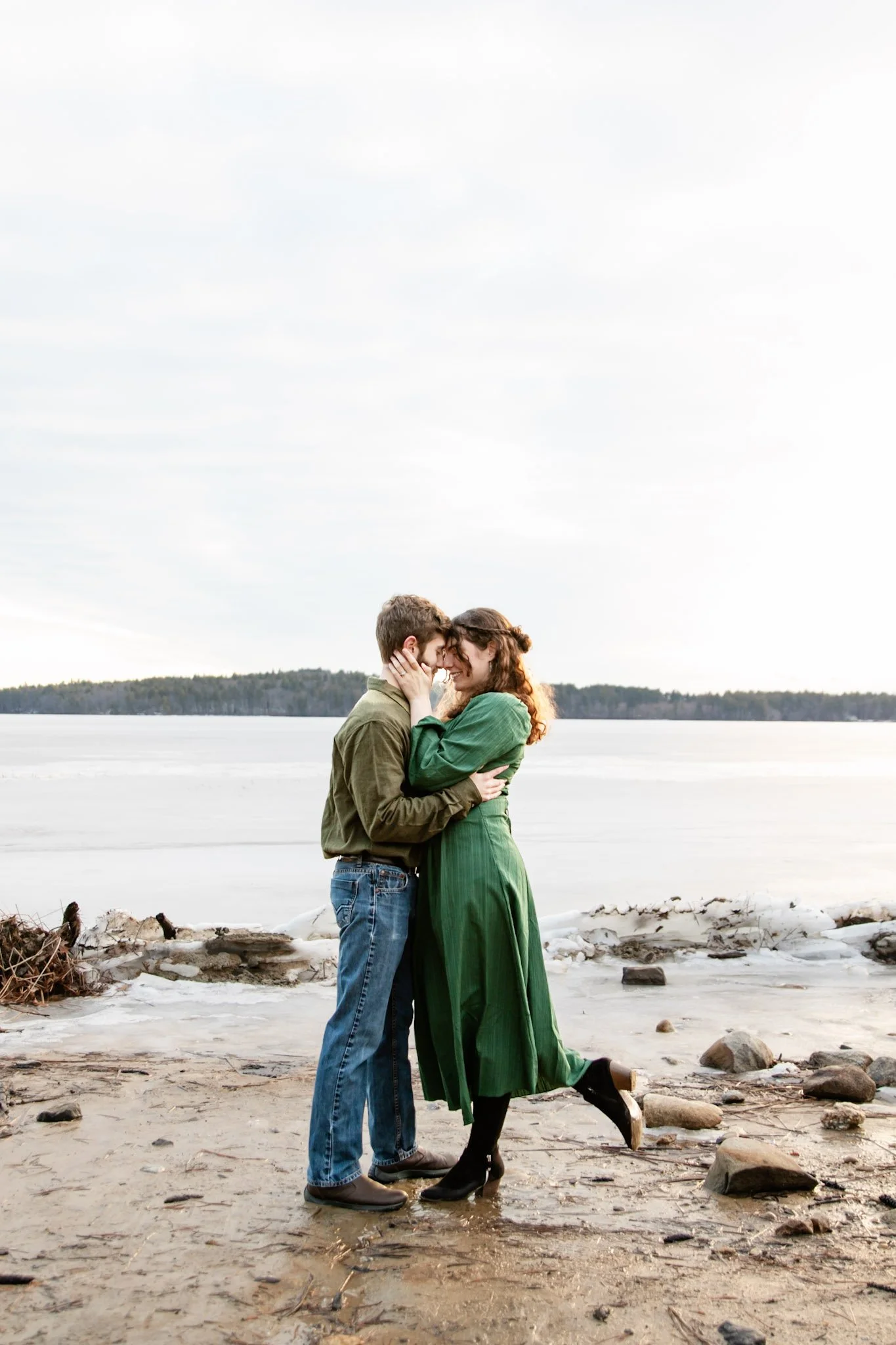 A couple stands close together on a beach by a lake, embracing and smiling at each other during daytime.