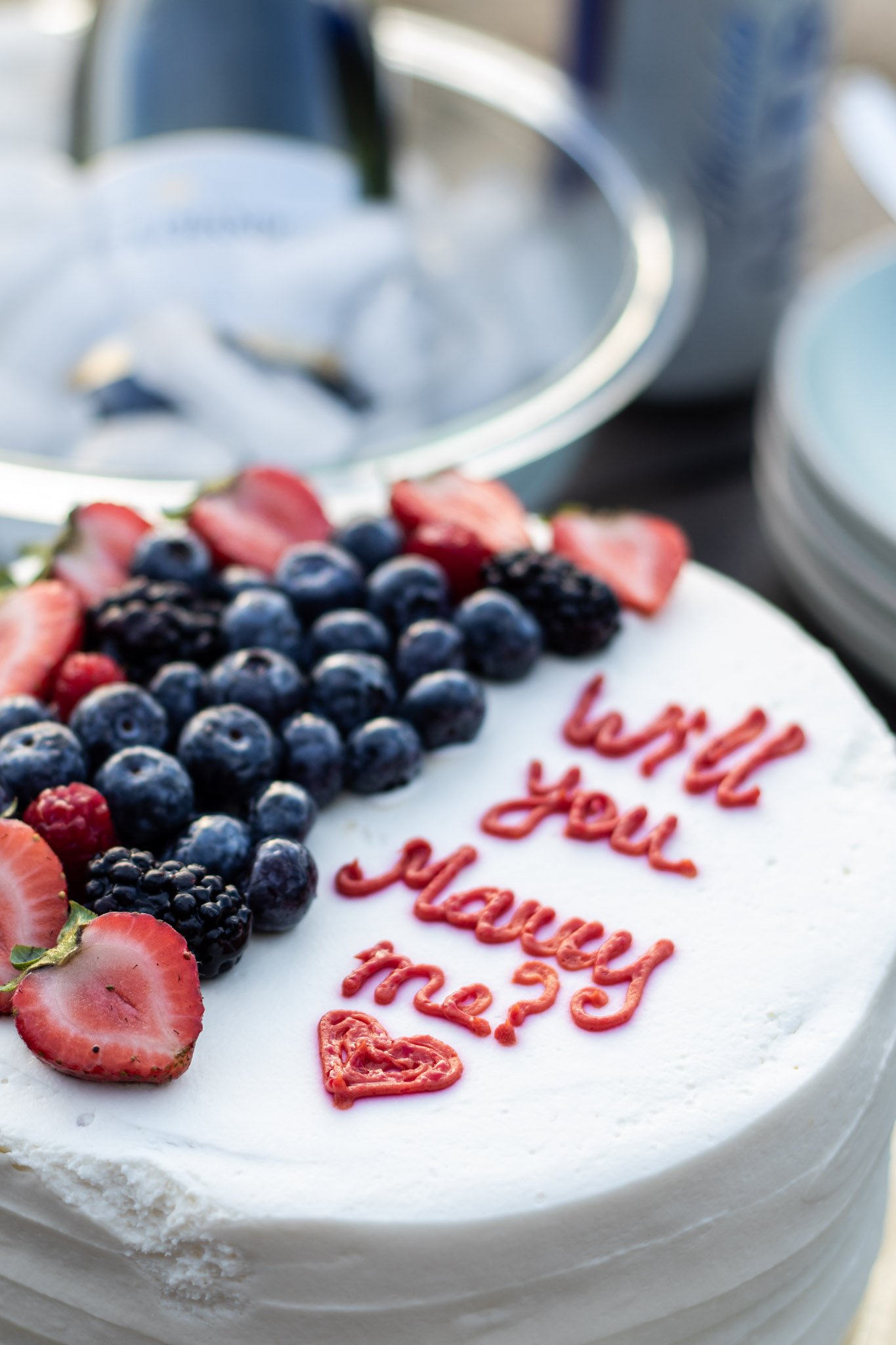 A birthday cake decorated with fresh berries and a message written in red icing reading 'will you marry me?'