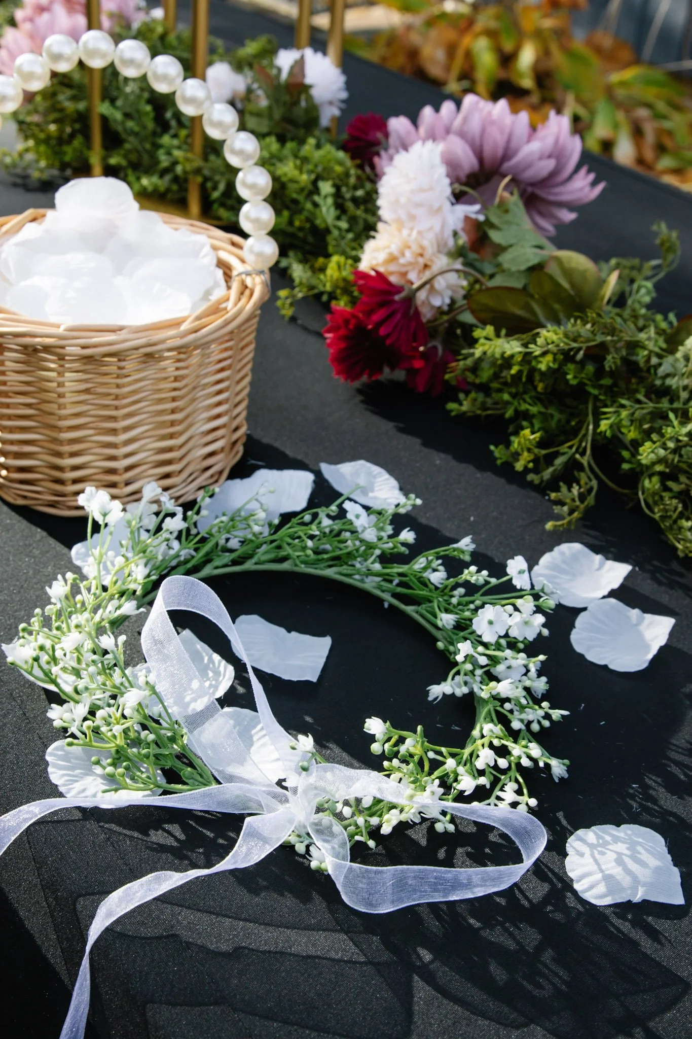 A floral wreath decorated with small white flowers and a white ribbon, laid on a black table. In the background, there are colorful flowers and a basket with a pearl handle.